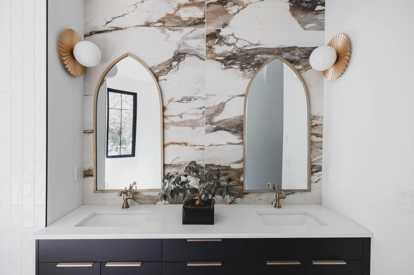 Modern bathroom vanity with two mirrors above sinks, a potted plant in between, and decorative wall sconces on both sides, featuring white globes and gold fan-like bases, set against a marble wall background.