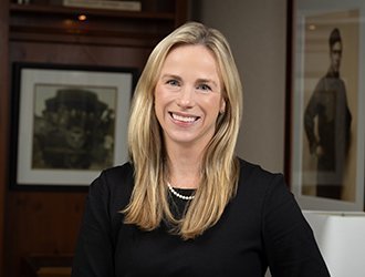 Portrait of a smiling woman with long blonde hair, wearing a black top and pearl necklace, in an office setting.