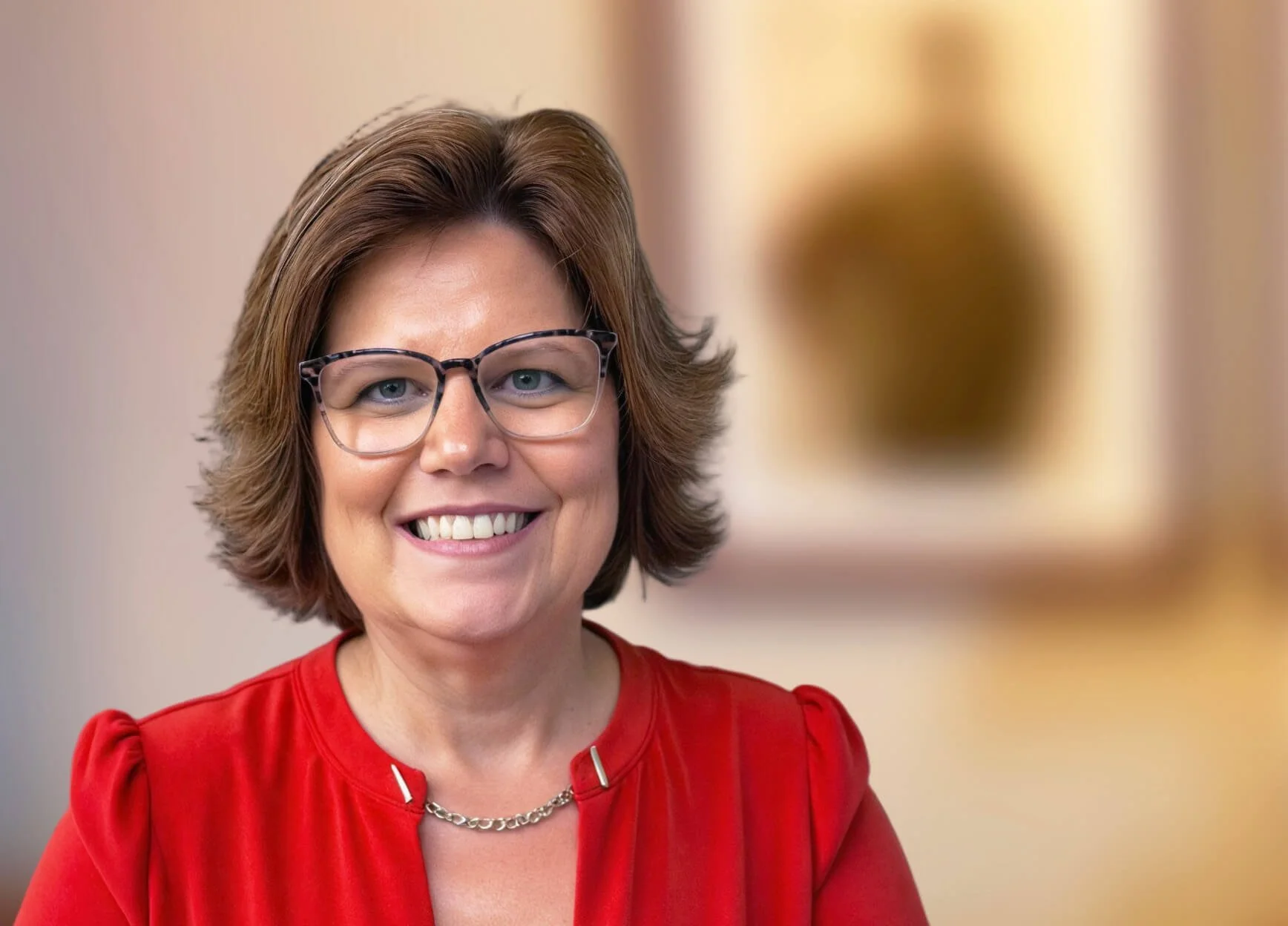 A smiling woman with short brown hair, wearing glasses and a red blouse, standing indoors with a blurred background.