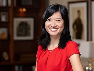 A woman with black hair, smiling, wearing a red top inside a room with framed pictures on wooden walls.