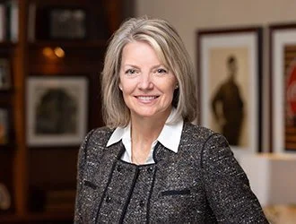 A smiling middle-aged woman with blonde hair, wearing a dark blazer over a white shirt, standing in an art gallery or office with framed artworks on the wall behind her.