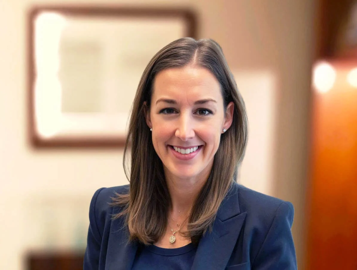 A woman with shoulder-length brown hair smiling, wearing a navy blazer and necklace, in an indoor setting with blurred background.