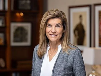 A smiling woman with long hair wearing a gray blazer and white blouse, standing in a room with framed artwork.