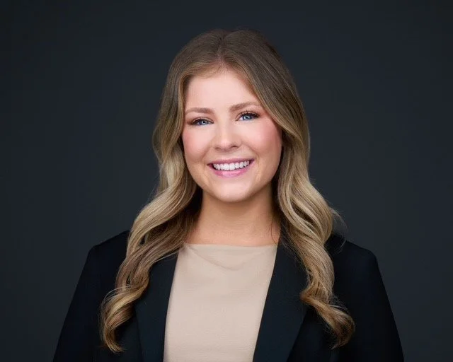 A young woman with long, wavy blonde hair, smiling, wearing a beige top and black blazer against a dark background.