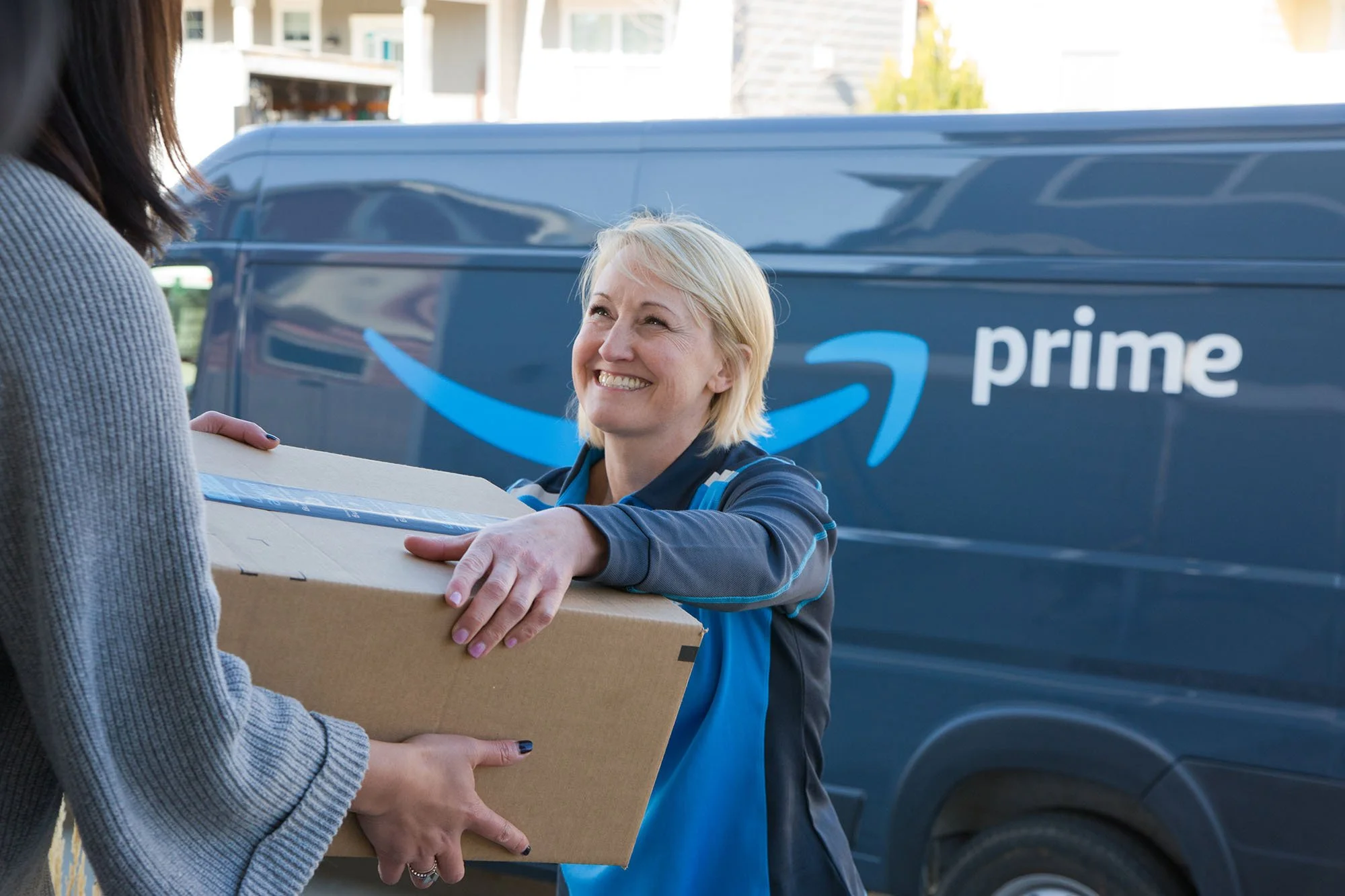 Amazon delivery person handing a package to a customer outside with an Amazon Prime delivery van in the background.