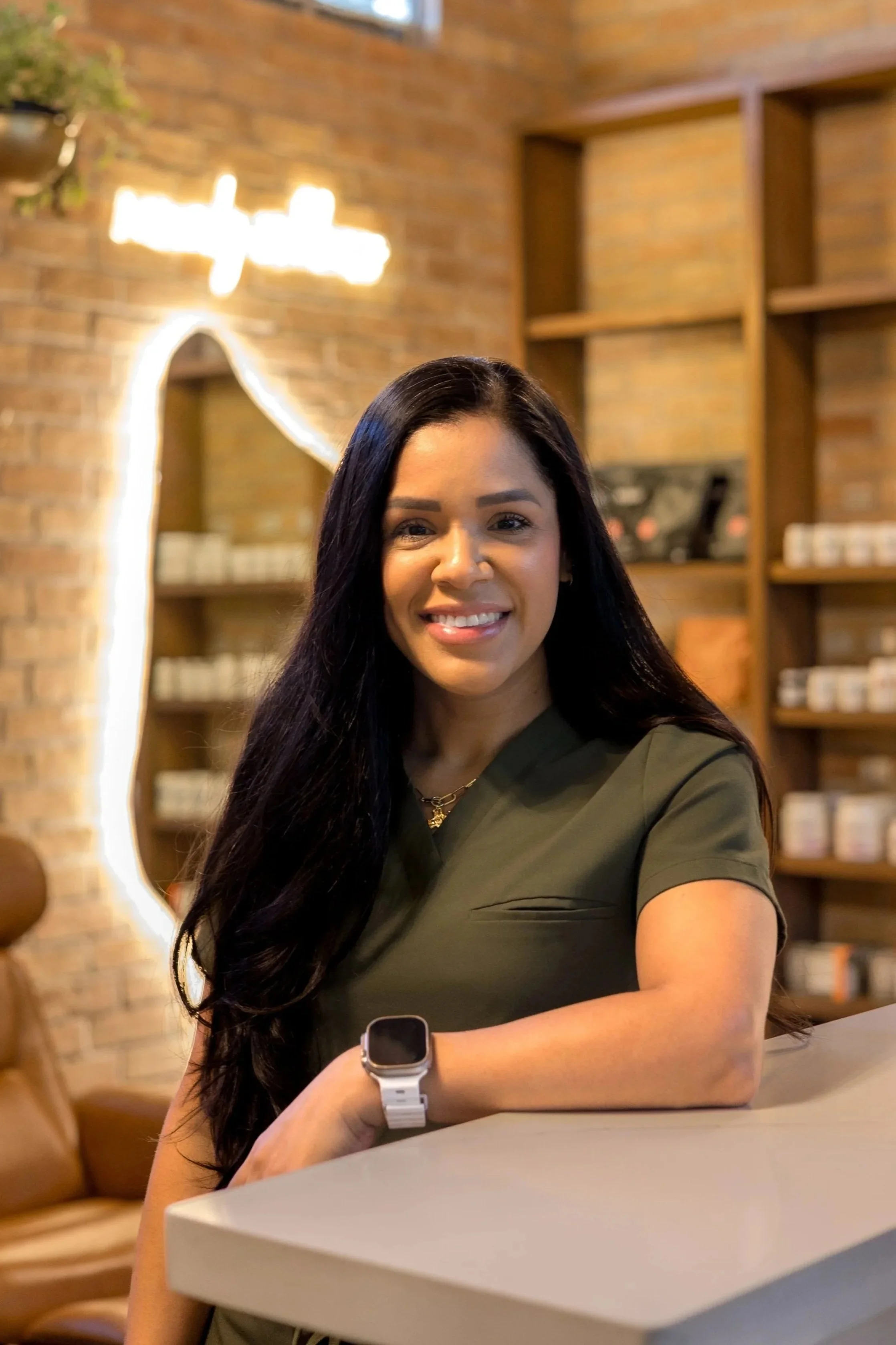 A young woman with long dark hair smiling and wearing a green shirt, sitting at a table in a cafe. She has a smartwatch on her left wrist and is looking at the camera. The background features a brick wall, wooden shelves, and decorative lighting.