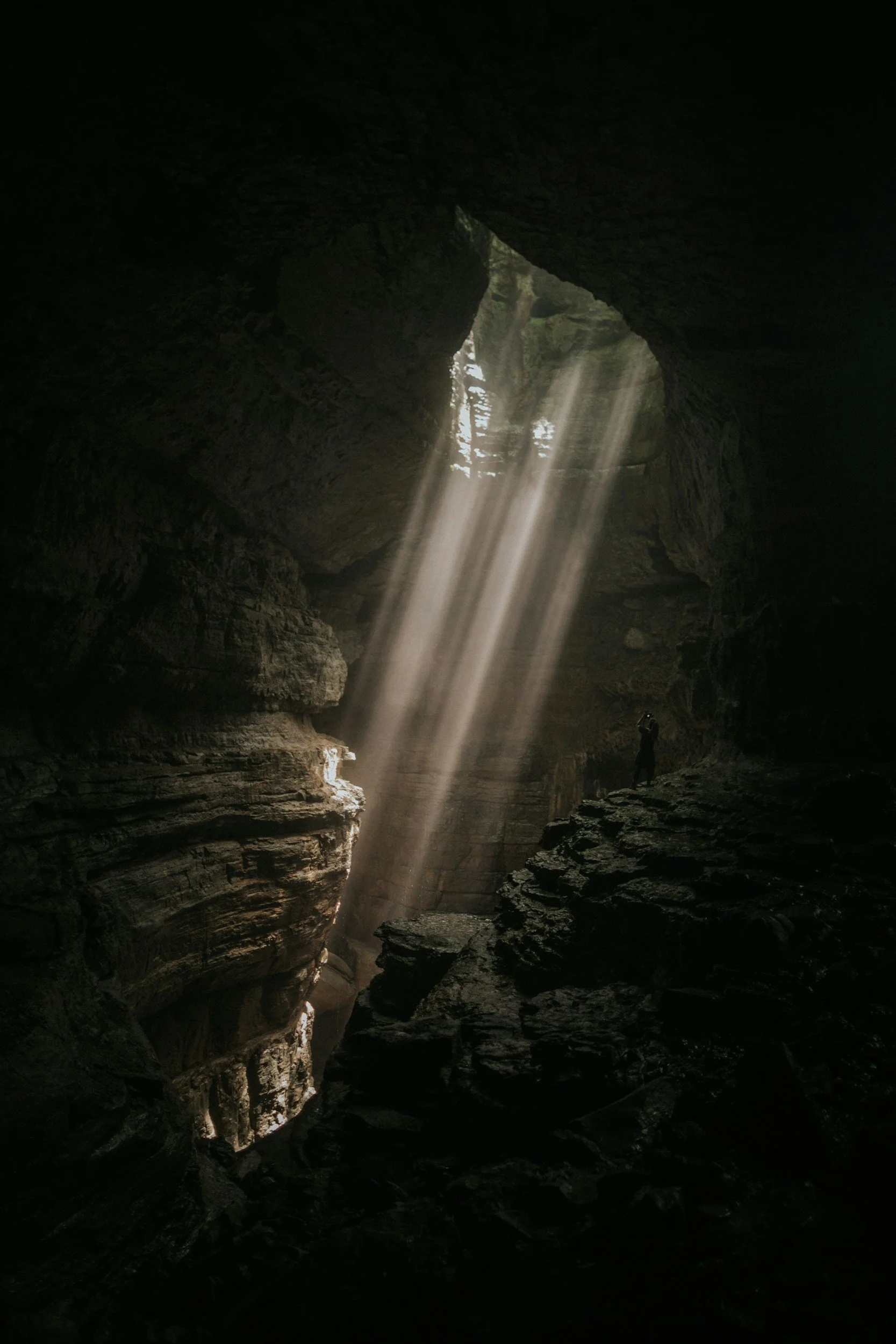 Sunlight streams through a cave opening, illuminating the dark interior and a person standing on a rocky ledge.