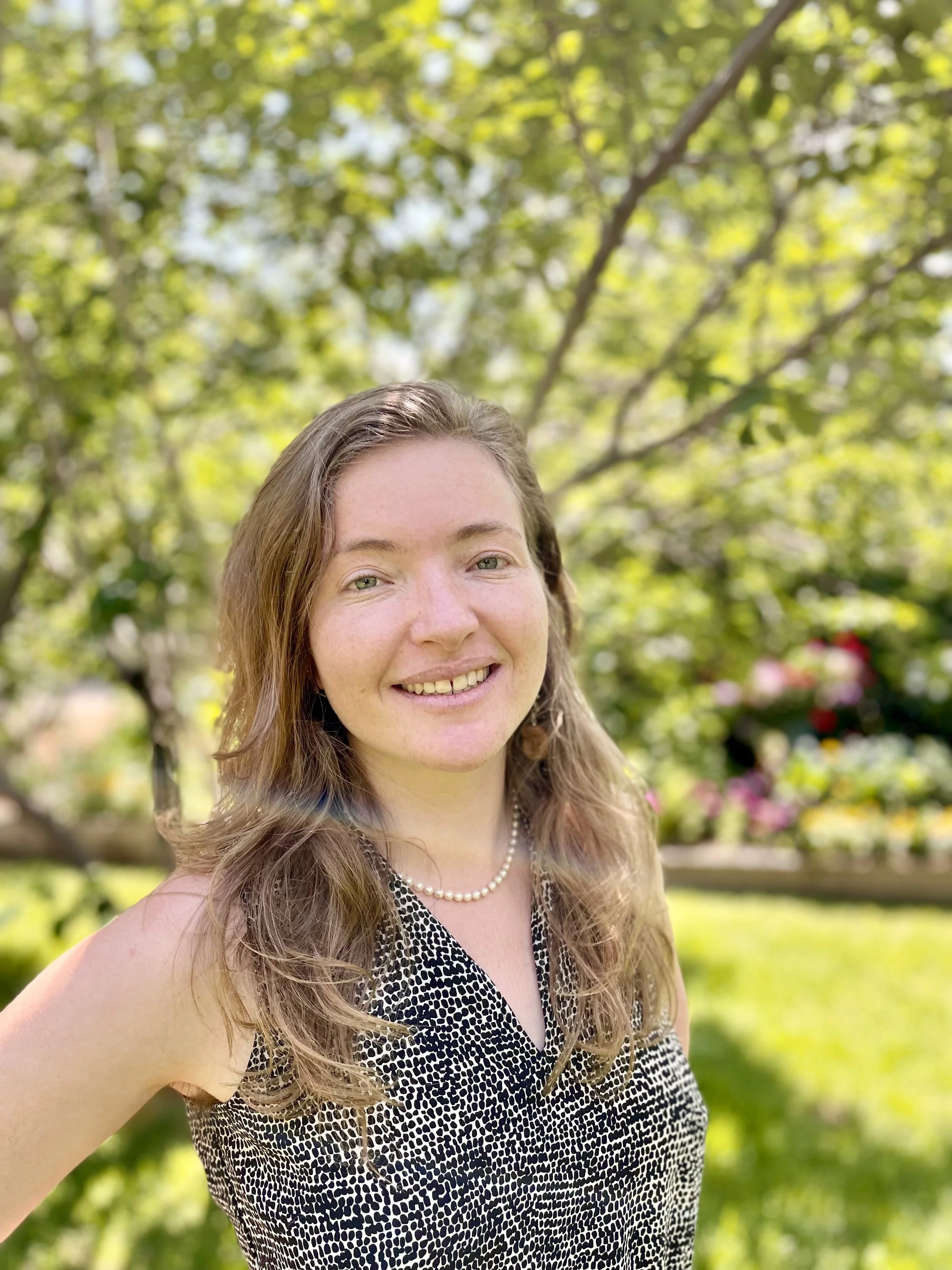 A young woman with long, wavy brown hair smiling outdoors, wearing a sleeveless black and white patterned dress and a pearl necklace, with a background of green trees and sunlight.