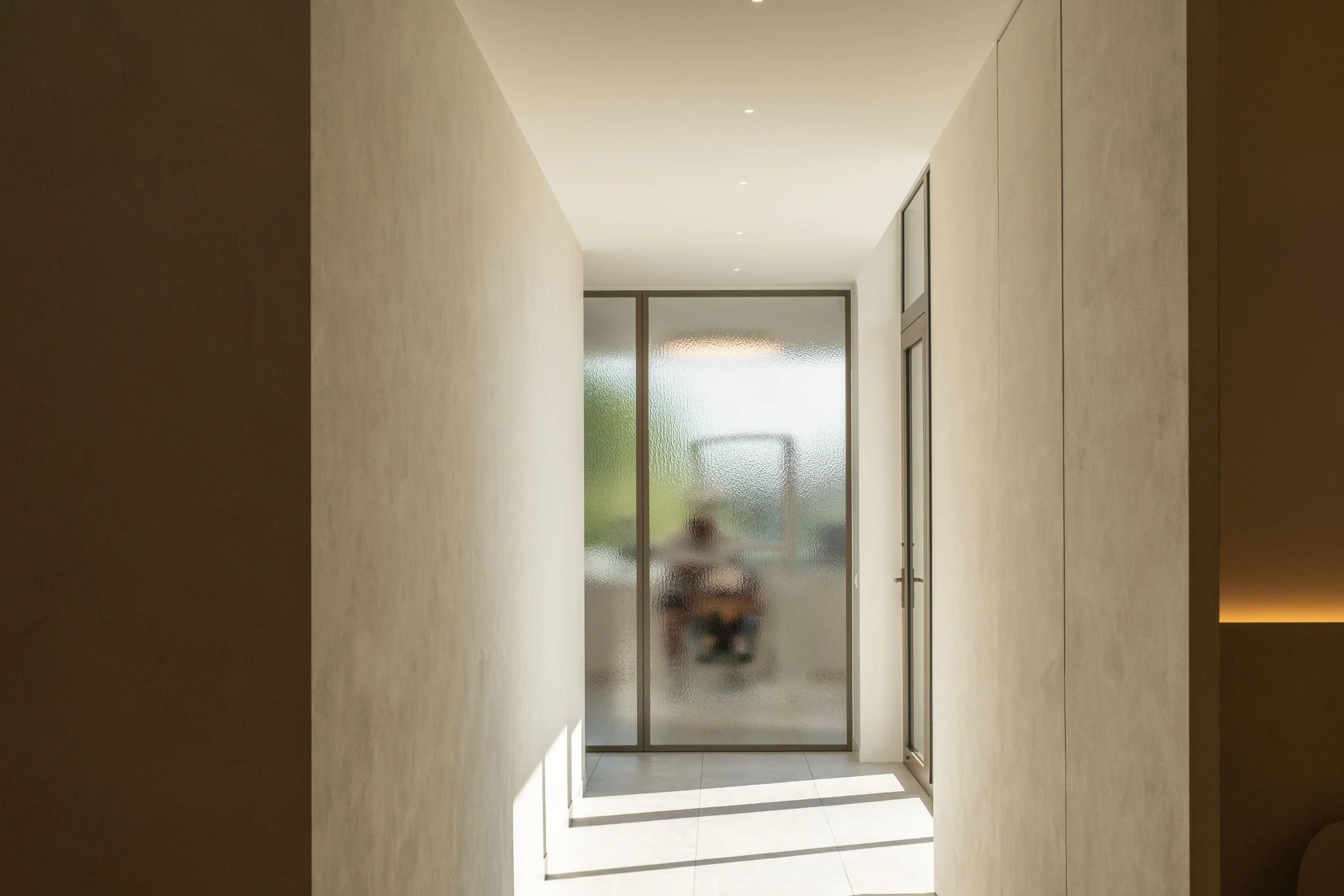 Een smalle, strakke gang met beige muren leidt naar een wand in figuurglas waarachter het silhouet van een werkplek zichtbaar is