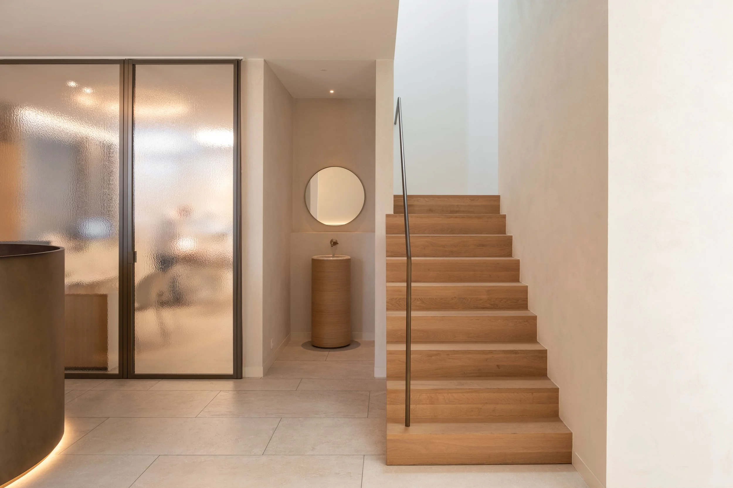 Een stijlvol en minimalistisch interieur met een houten trap, een glazen wand met matglas en een nis met een cilindrische wastafel en ronde spiegel