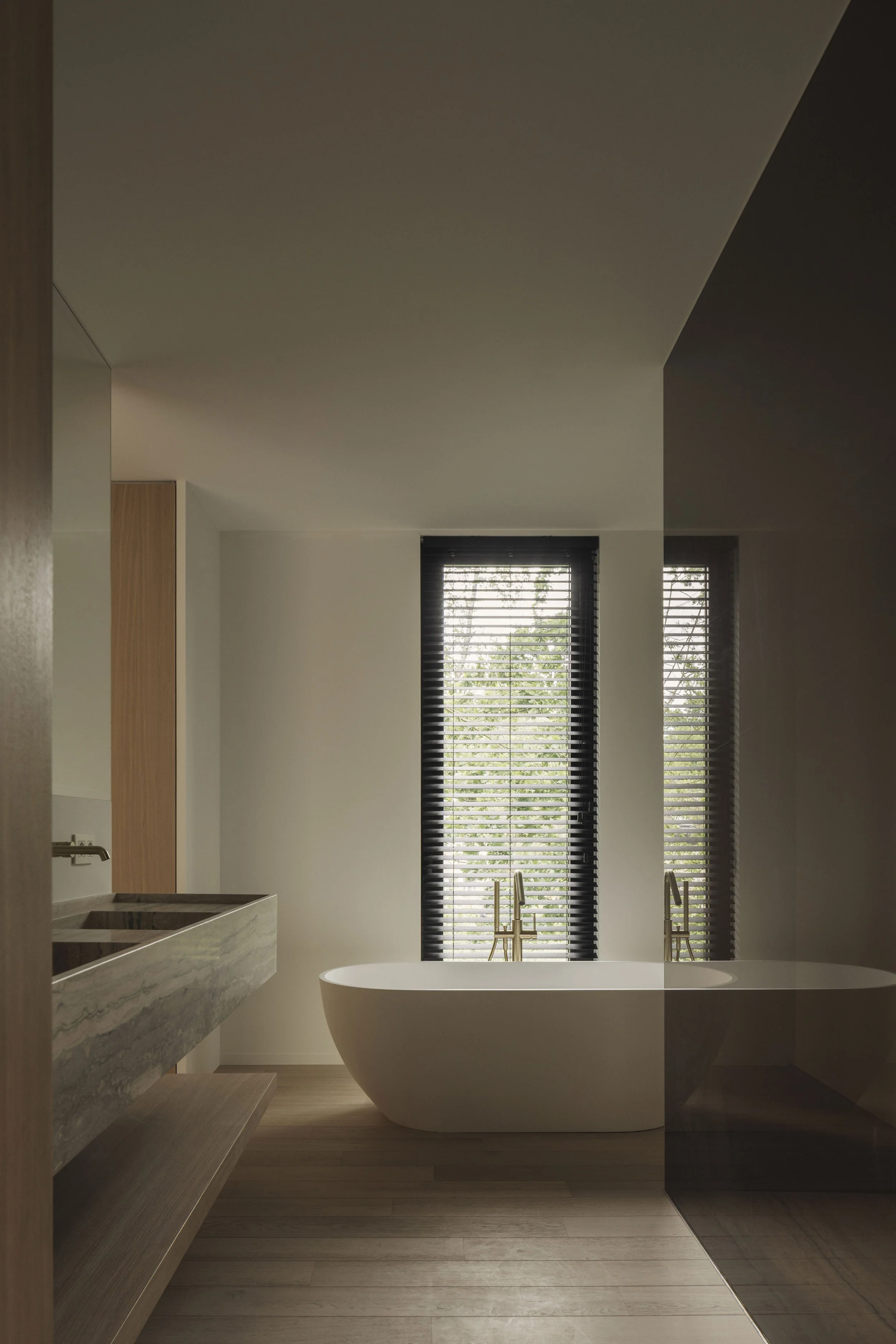 minimalistische en luxueuze badkamer met een vrijstaand bad, marmeren wastafel, houten details en een raam met lamellen dat uitzicht biedt op een groene omgeving