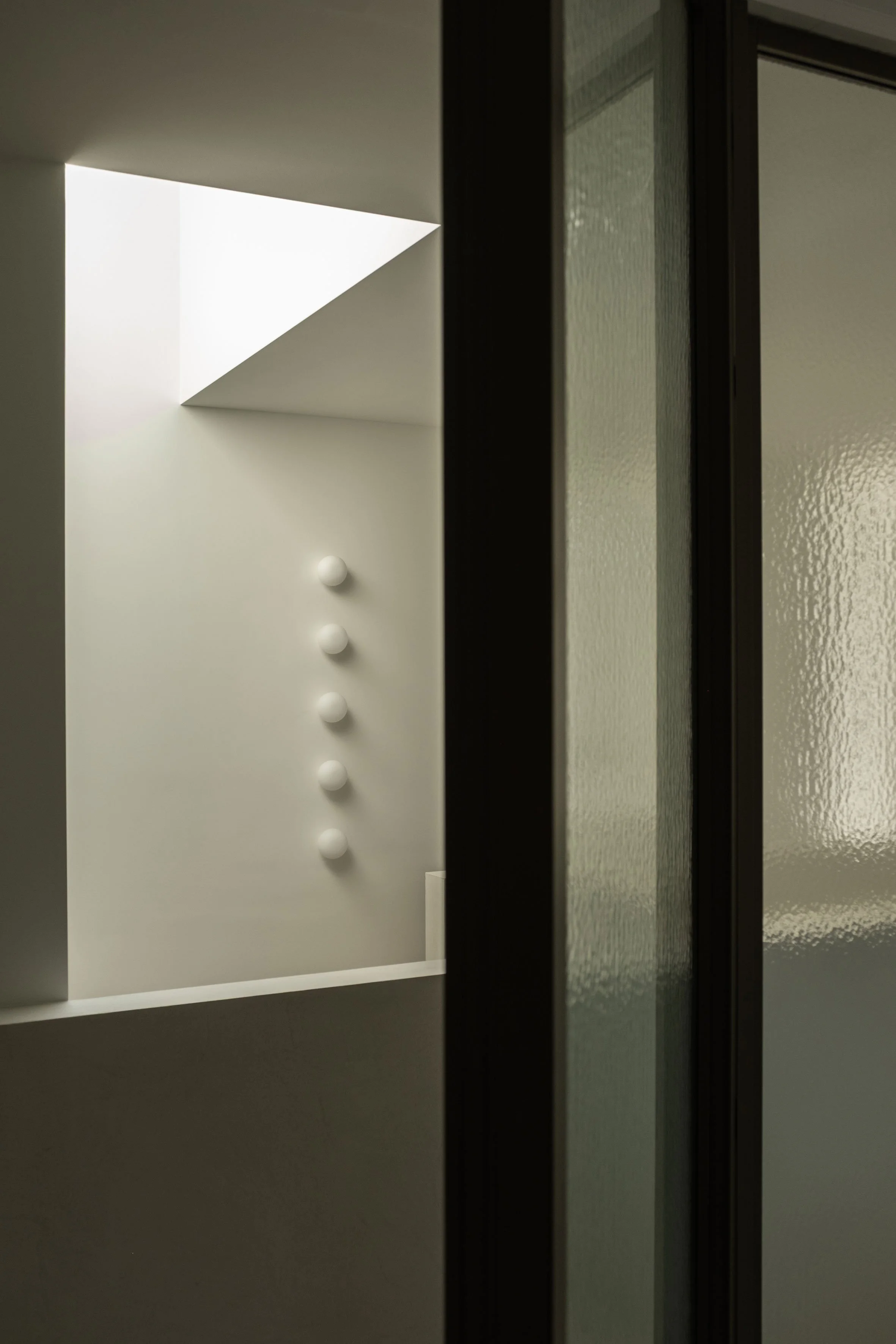 Een doorkijk door een glazen deur naar een minimalistische, strakke ruimte met een lichtkoepel en een rij bolvormige wandlampen op een witte muur