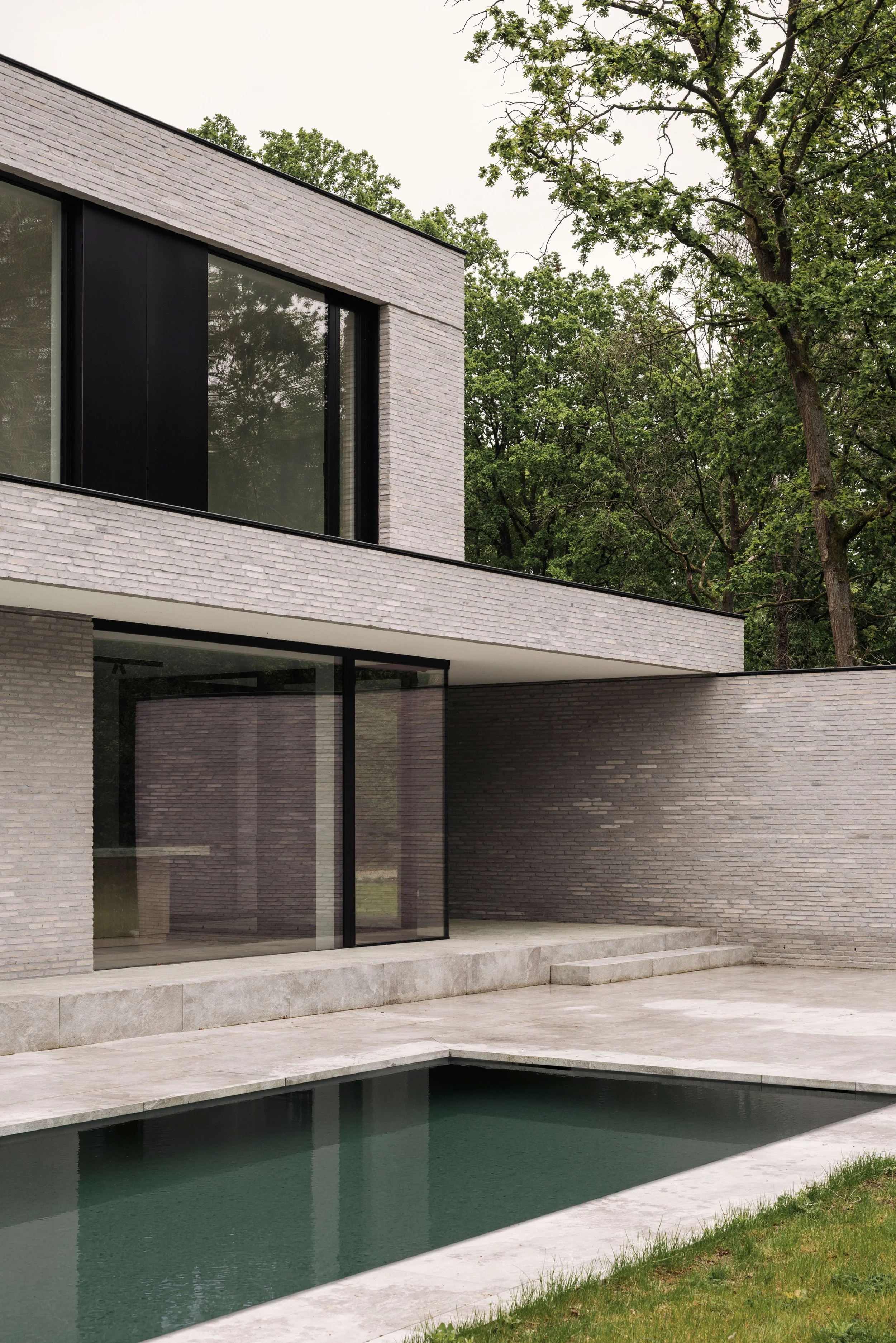 moderne, minimalistische woning met een grijze bakstenen gevel, grote raampartijen en een zwembad in een bosrijke omgeving