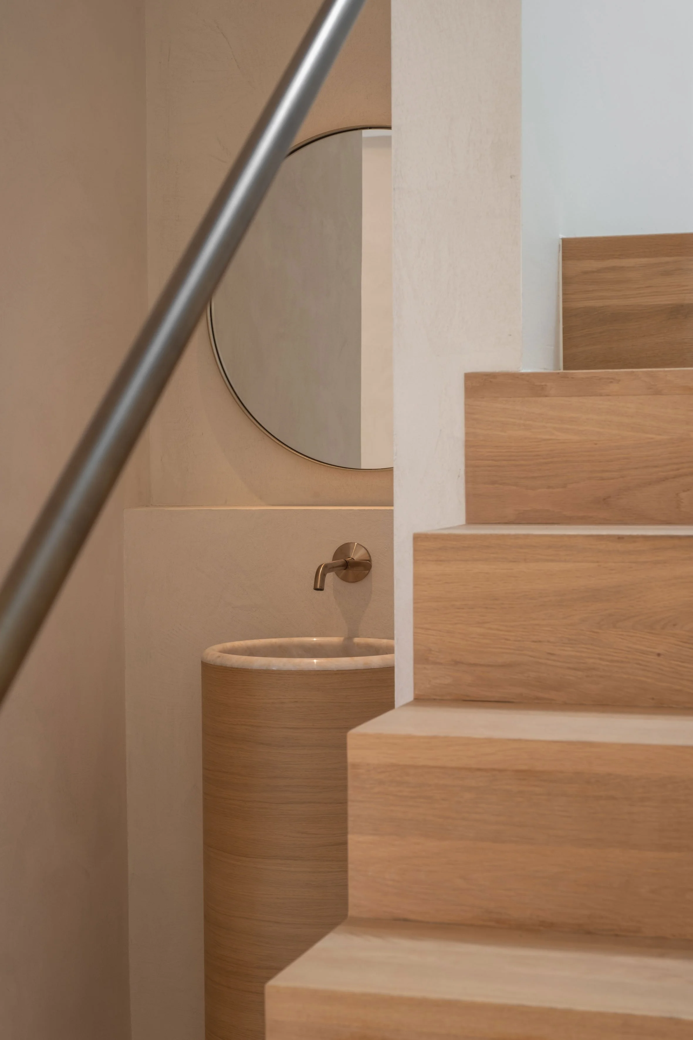 Een detailopname van een minimalistisch interieur met houten traptreden en een nis waarin een vrijstaande cilindrische wastafel met een ronde spiegel staat
