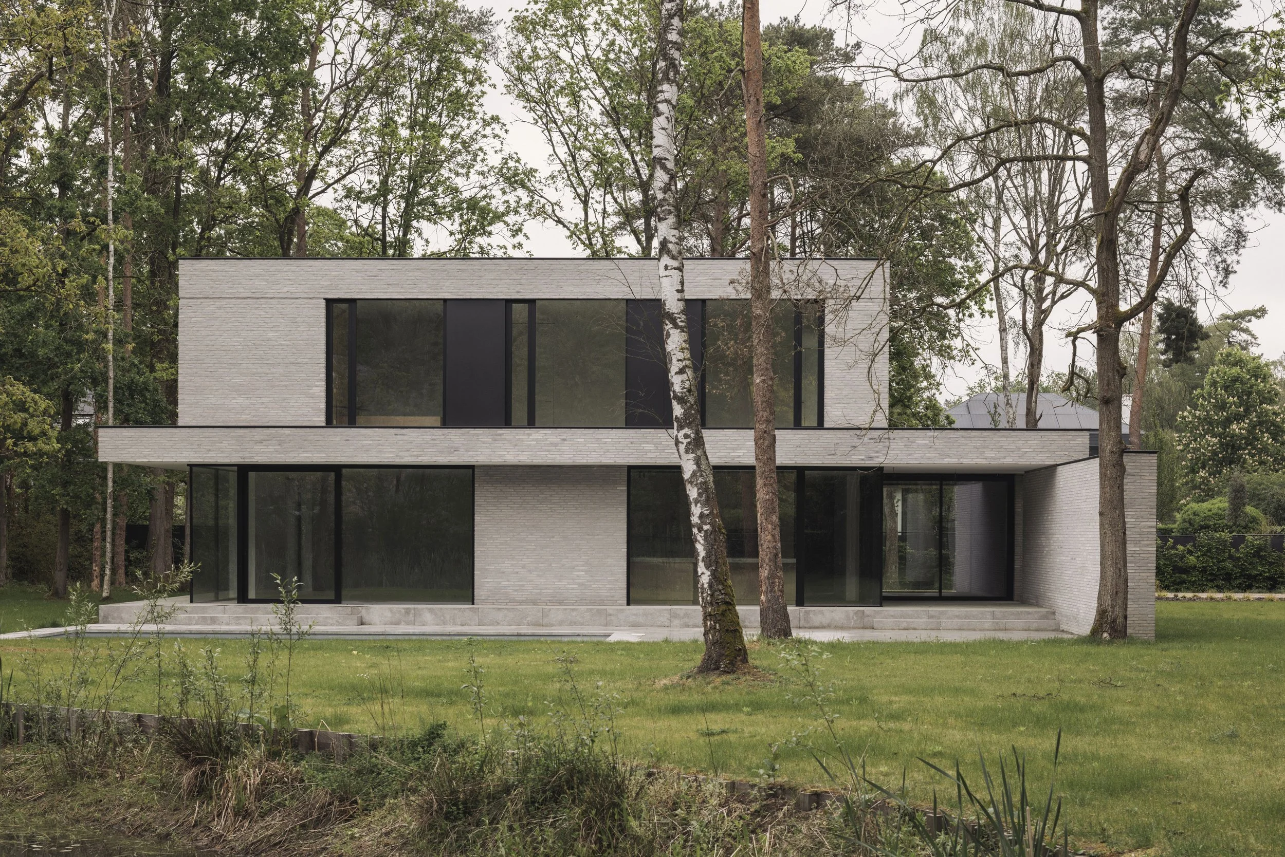 moderne, minimalistische villa in lichtgrijze baksteen met twee verdiepingen, gesitueerd in een bosrijke omgeving. Strak zwart aluminium buitenschrijnwerk