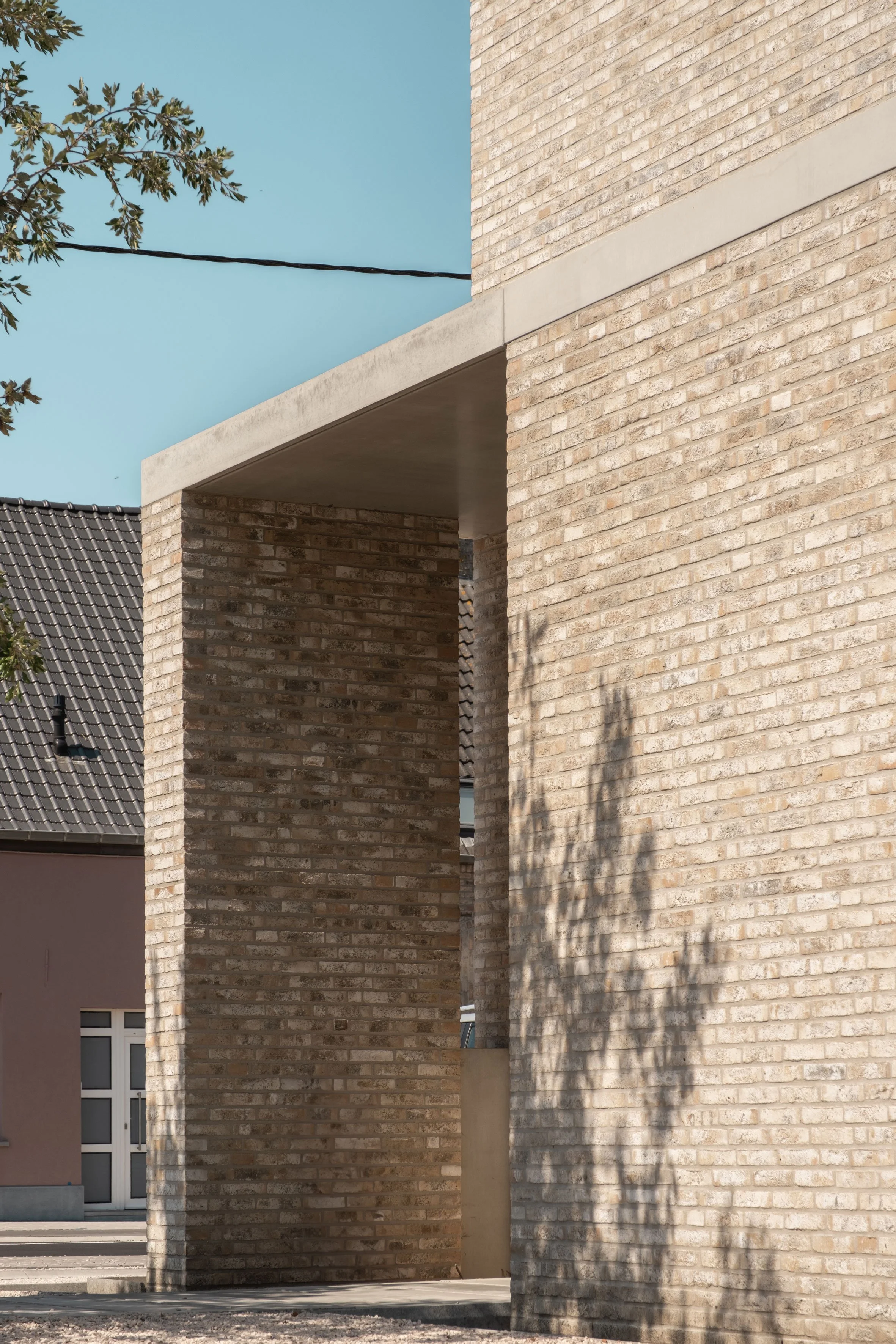 Een detailopname van een modern bakstenen gebouw met een strakke betonnen overkapping en een boomdenschaduw die op de lichtgekleurde gevel valt.