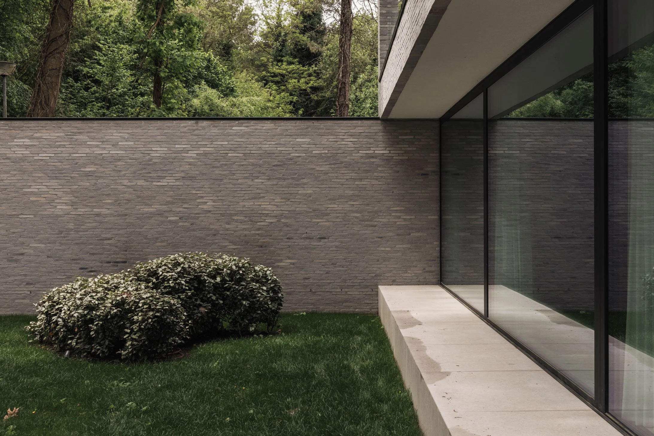 Een strakke glazen gevel van een moderne woning met zicht op een grijs gemetselde tuinmuur, een groen gazon met struiken en bomen op de achtergrond.