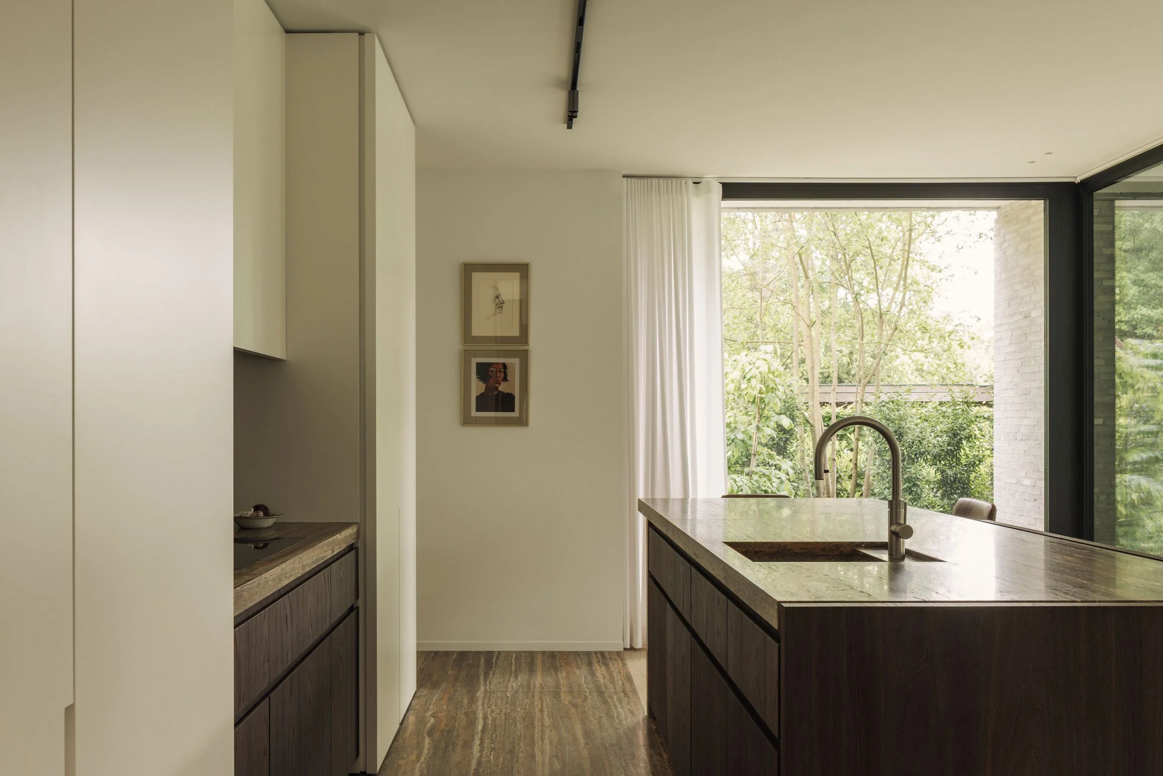 Overzichtsfoto van een strakke, moderne keuken toont een kookeiland met een adergewelfd natuurstenen werkblad en een RVS-kraan, gecombineerd met donkerhouten kasten en een uitzicht op de groende tuin in de achtergrond