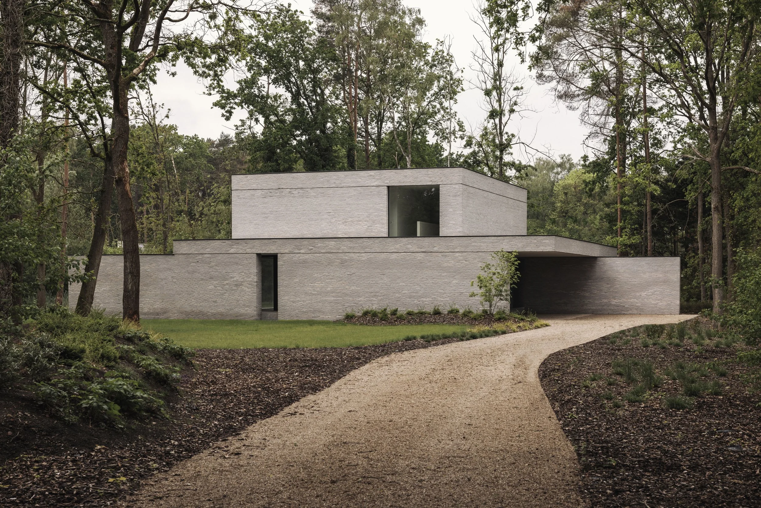 moderne, minimalistische woning met een grijze genuanceerde bakstenen gevel, grote raampartijen en een overdekte inkom in een bosrijke omgeving