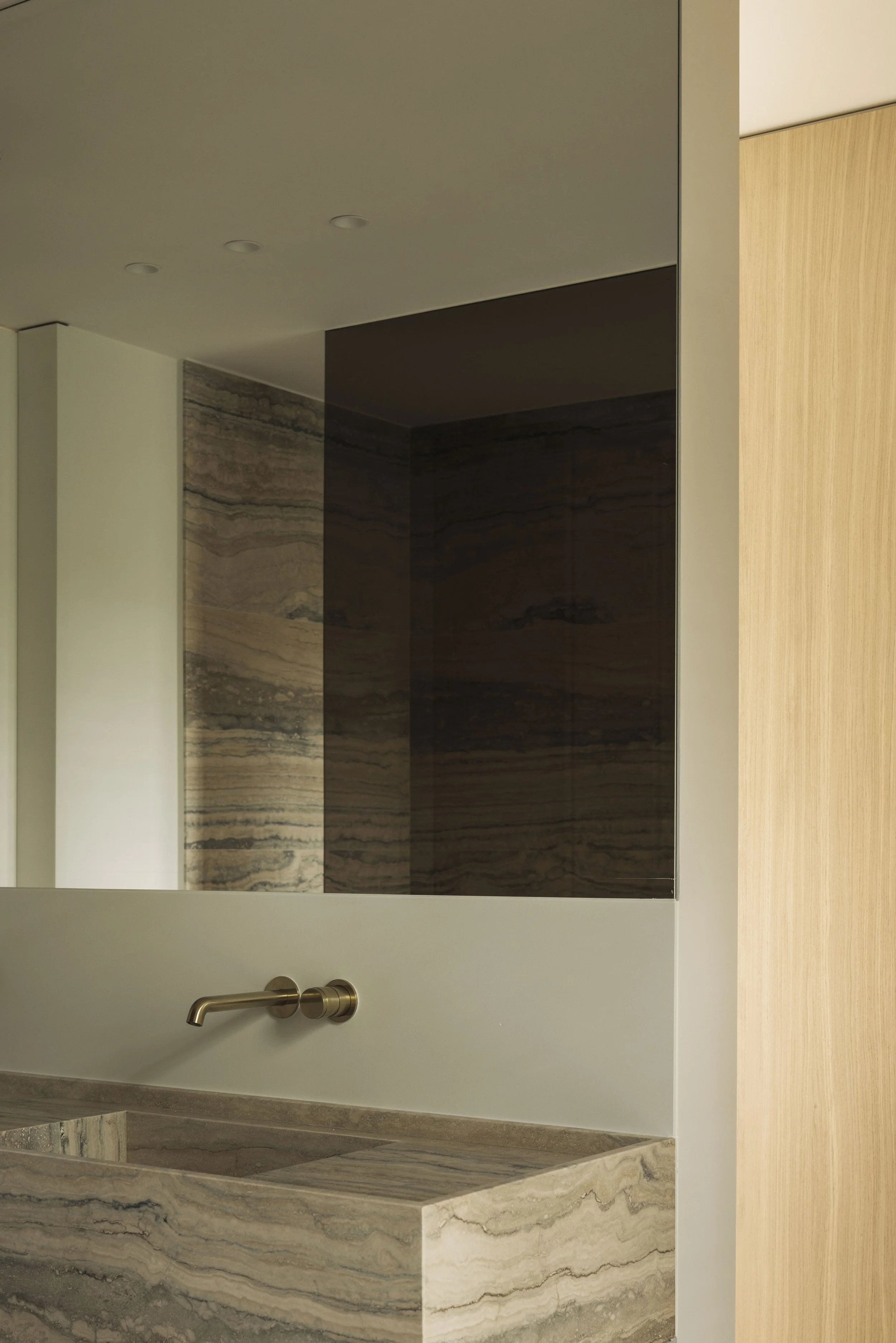 modern en luxueus badkamerinterieur met een opvallende beige travertin wastafel, een minimalistische gouden wandkraan en een grote spiegel die de travertin achterwand reflecteert.