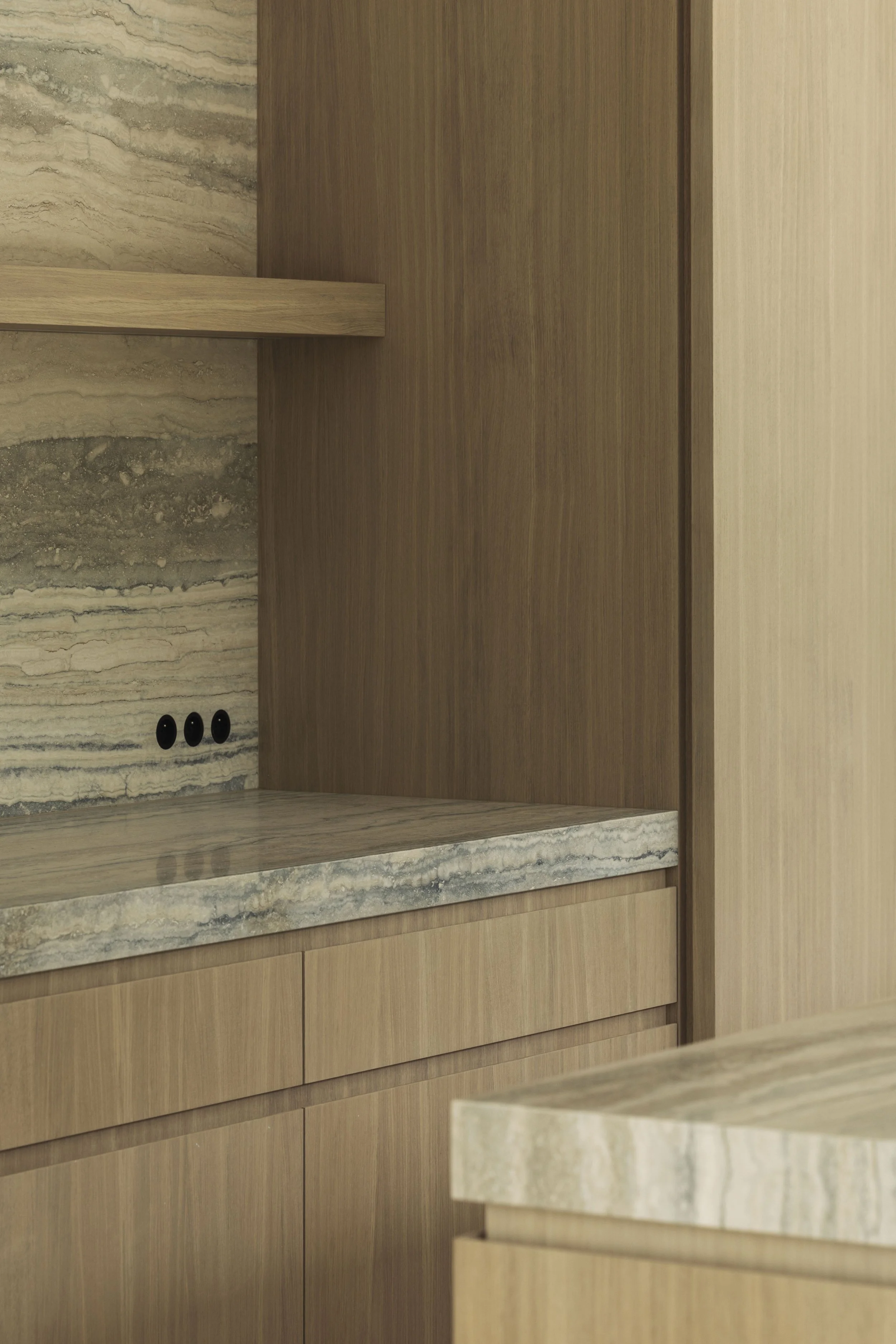 modern interieurontwerp toont een keuken met eikenhouten kasten en panelen, een opvallende travertin natuursteen in beige tinten voor aanrechtblad en achterwand met een bijpassende zwevende plank, en drie ingebouwde zwarte ronde stopcontacten.
