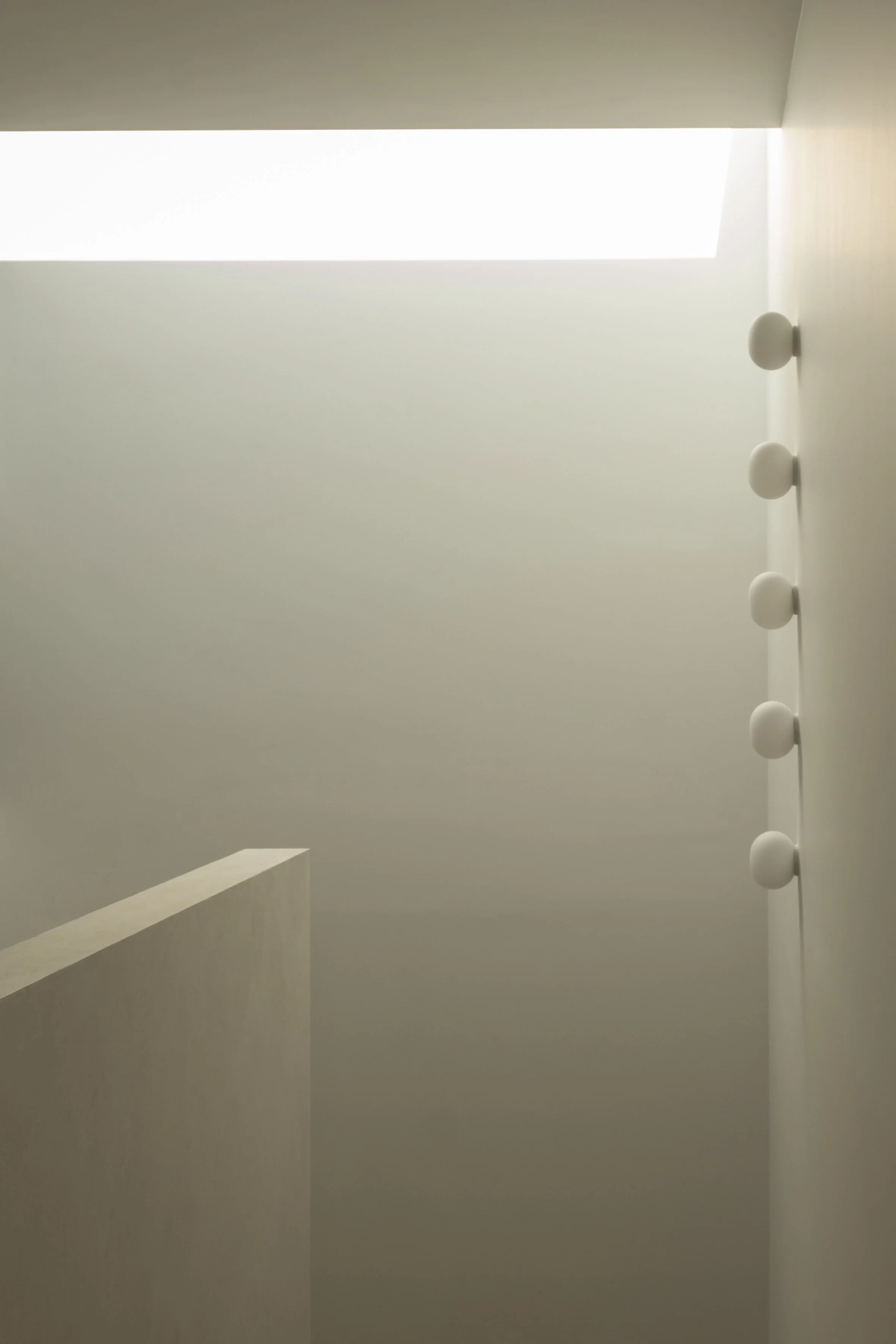 Een minimalistische binnenruimte met strakke beige muren, verlicht door een lichtkoepel en voorzien van een verticale rij bolvormige wandlampen