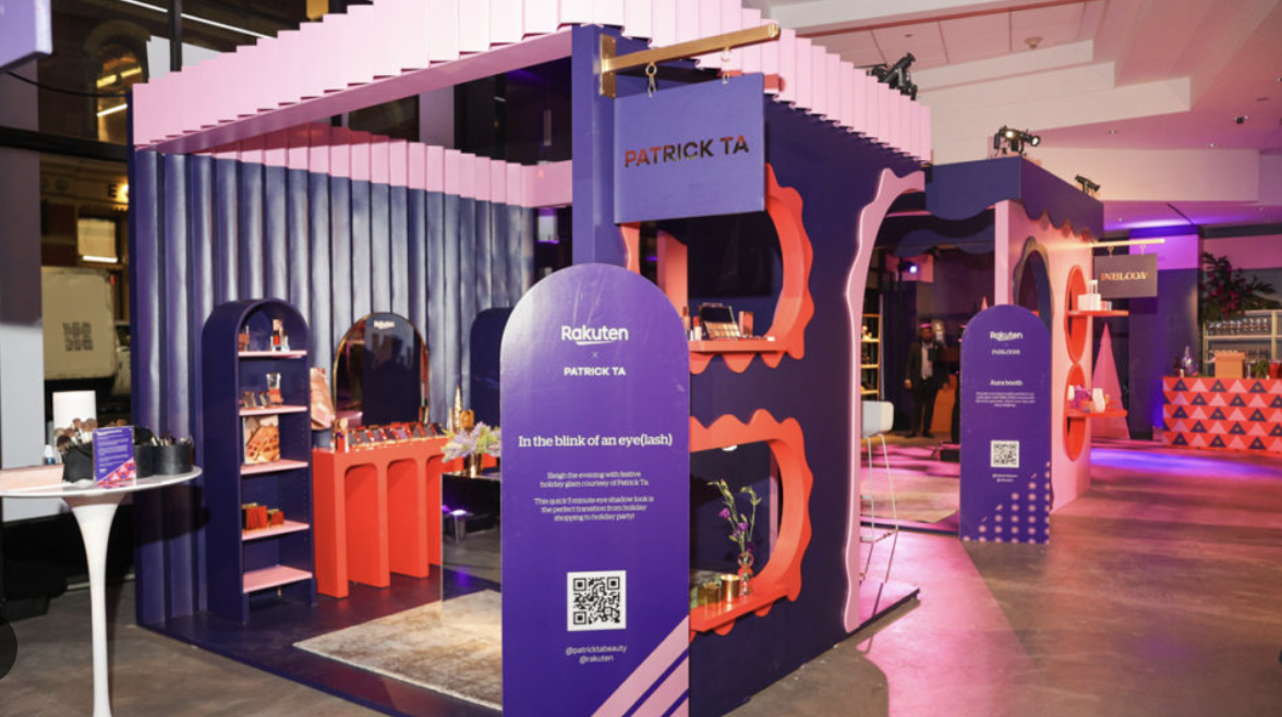 Colorful booth with purple and red accents at an event, showcasing Rakuten products and promotional materials, with a sign encouraging quick eye blink during a holiday glow experience.