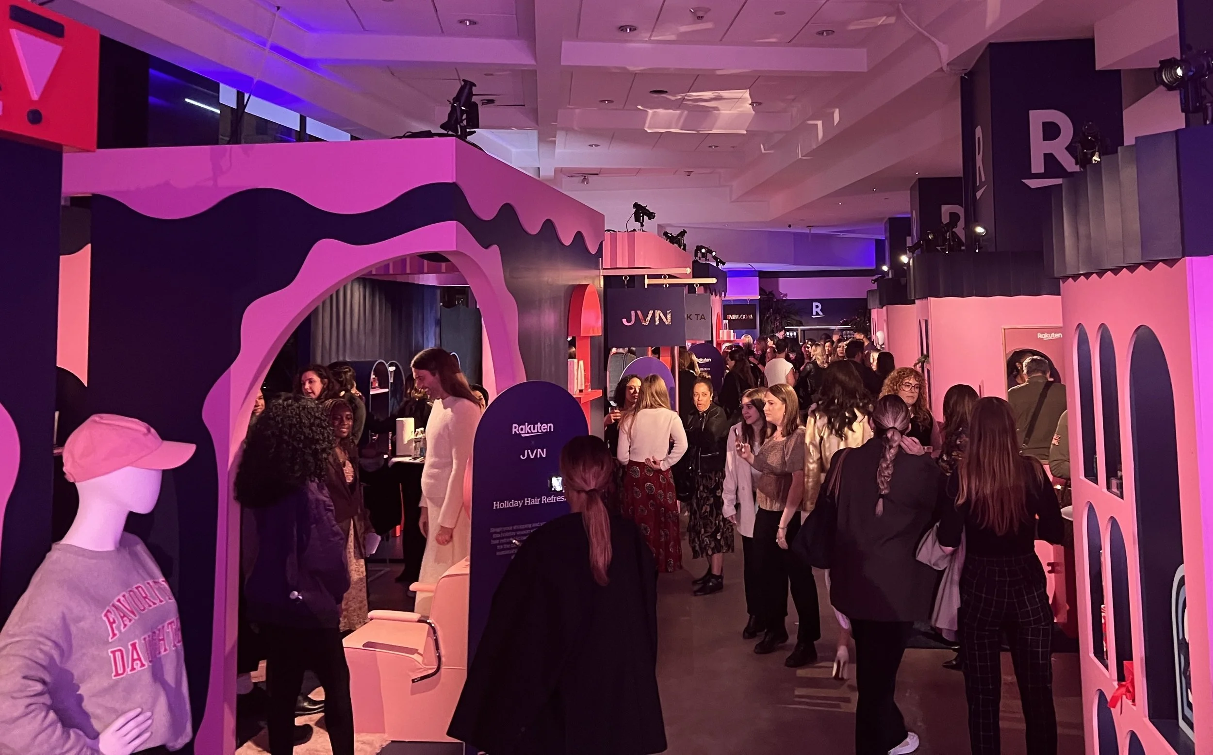 A busy indoor event with pink and black decor, mannequins, and numerous people engaging at various booths and displays.
