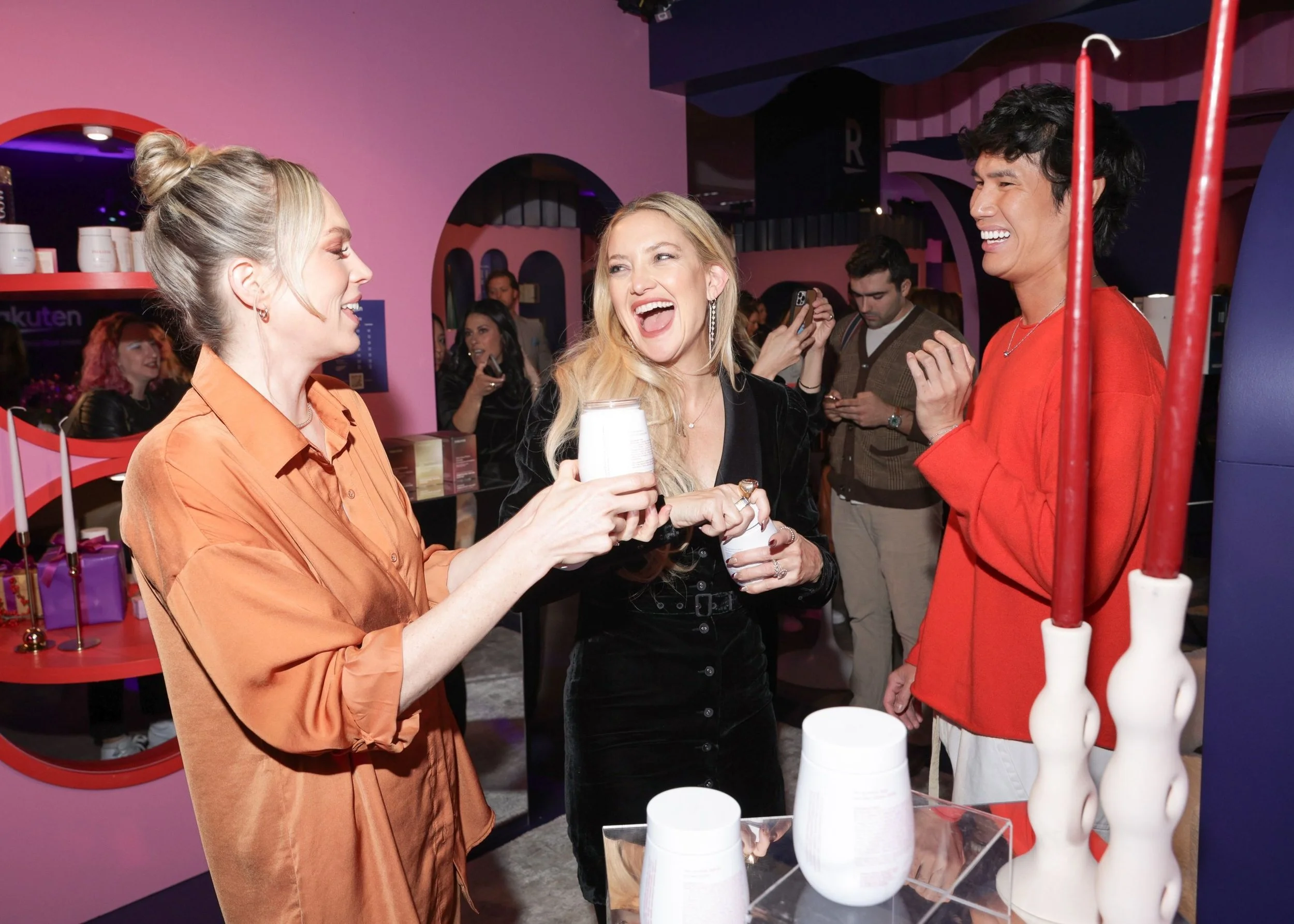 Three women engaging in conversation at a social event, smiling and holding drinks, with other people in the background. The setting features pink walls and modern decor.