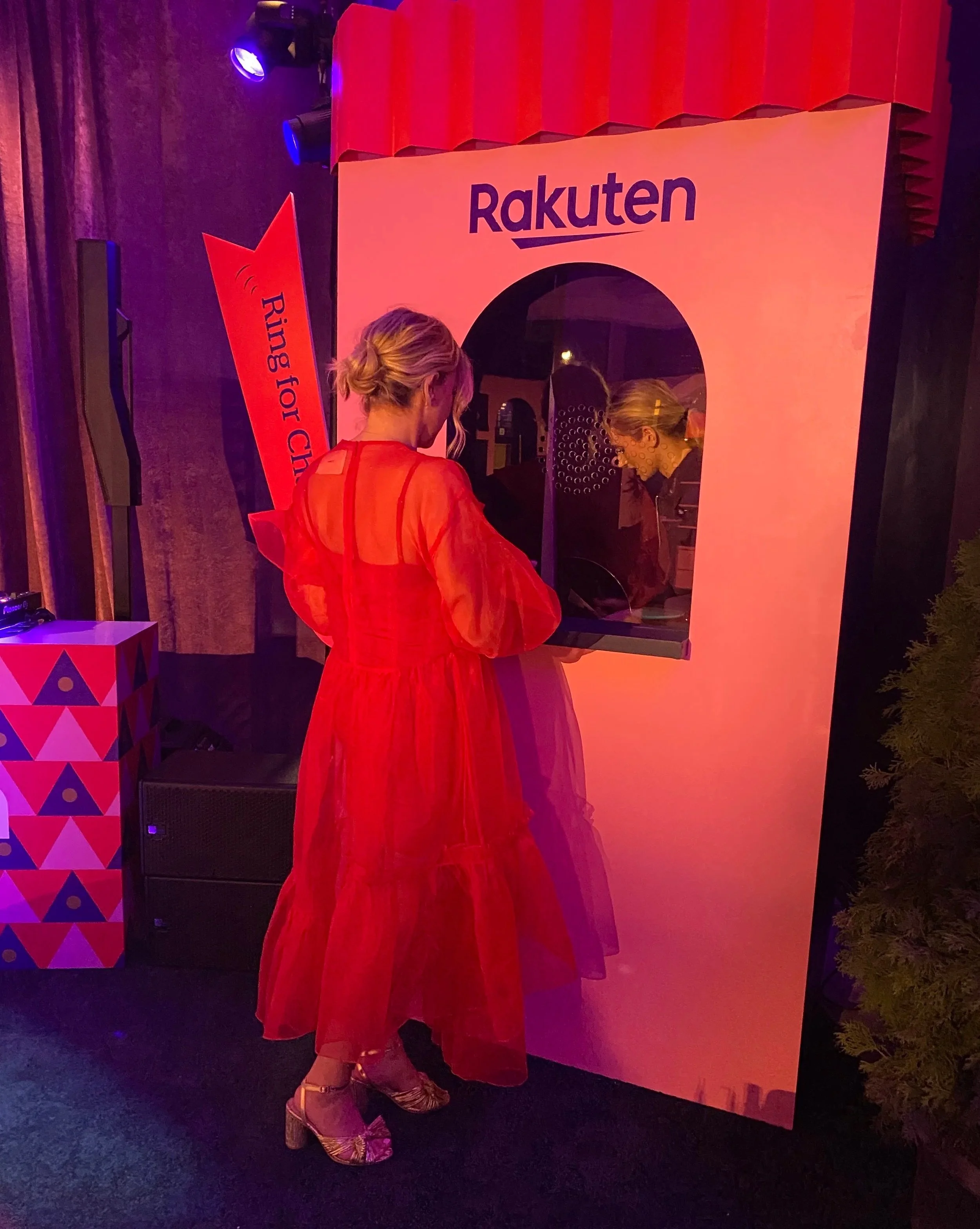 A woman in a red dress standing at a Rakuten-themed photo booth or kiosk with a mirror or screen in an indoor setting with colorful lighting.