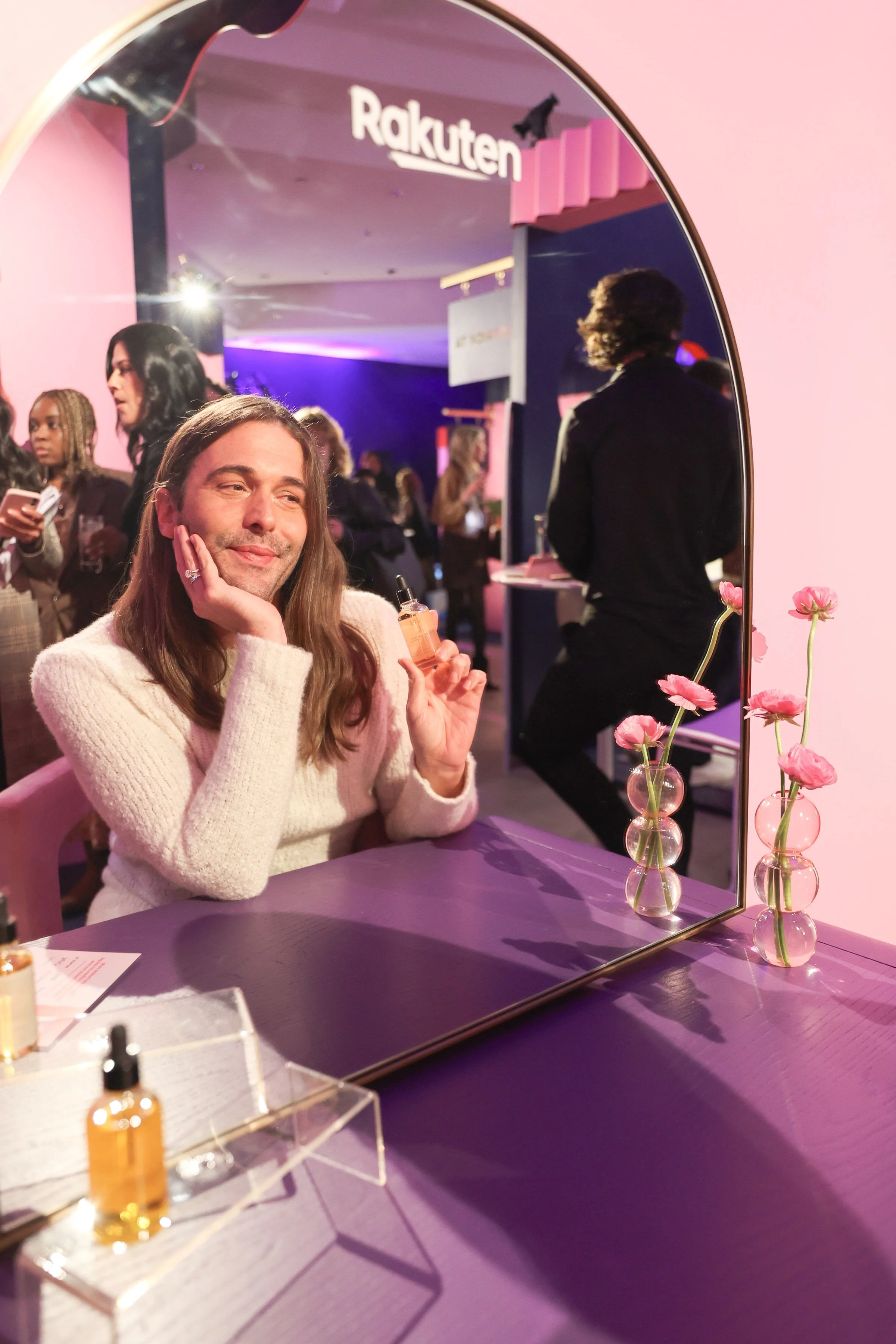 A woman sitting at a table in front of a mirror, holding a small bottle of skincare product, smiling with her hand resting on her face, in a brightly lit space with pink and purple lighting, with other people in the background and pink flowers in vases on the table.