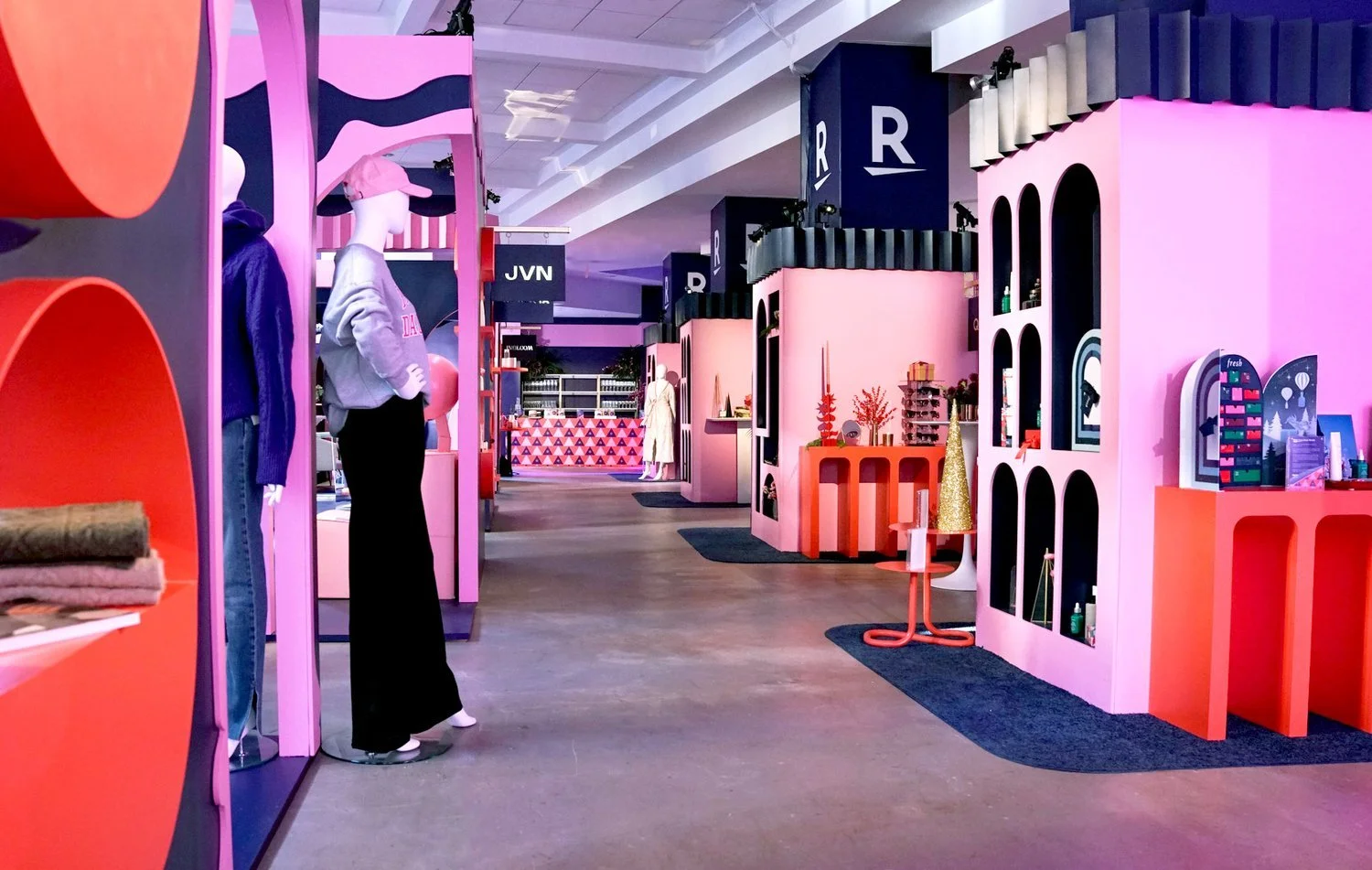 Colorful boutique store with pink, purple, blue, and black display units and mannequins dressed in casual clothing. The store has geometric and abstract decor, with shelves holding accessories and folded clothes.