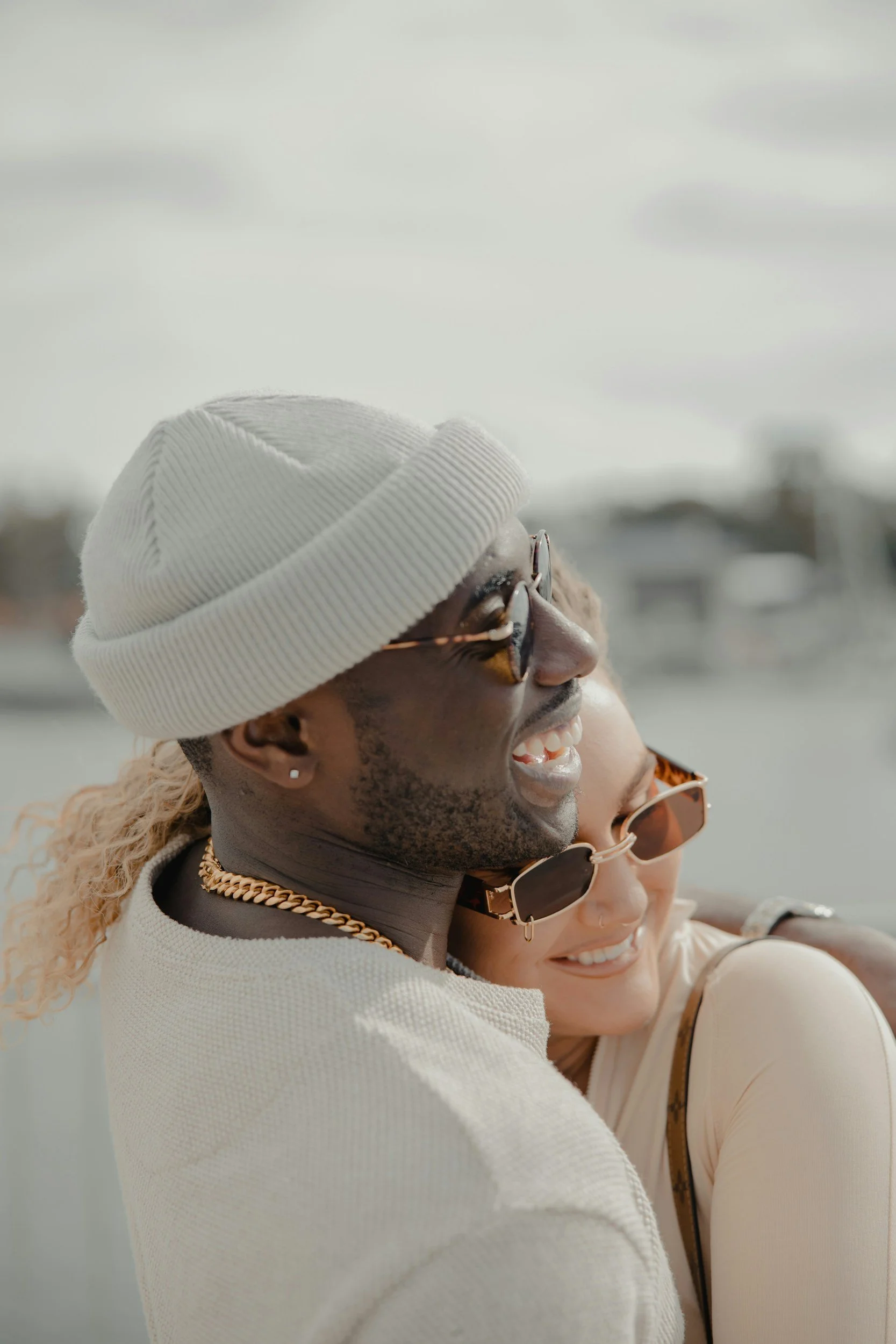 A man and woman smiling and hugging outdoors, wearing sunglasses, with the man wearing a white knit hat and necklace.