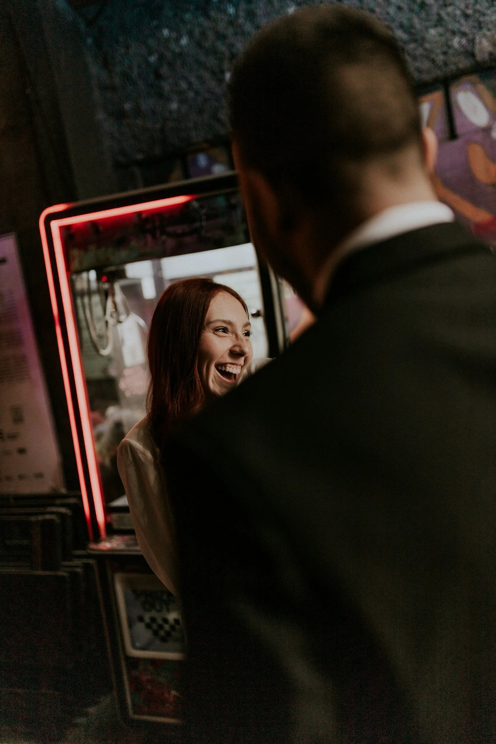 A woman smiling in front of a mirror, with a man partially visible from behind, inside a dimly lit room with various colorful posters and a neon sign.
