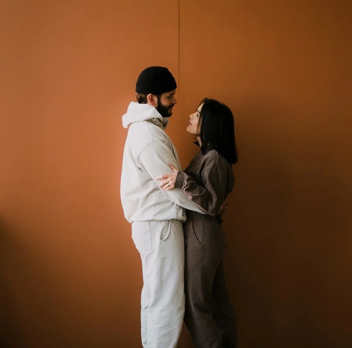 A man and woman standing close, facing each other, in front of a plain brown wall. The man has a beard, is wearing a black beanie, a light hoodie, and matching sweatpants. The woman has shoulder-length black hair, and is wearing a brownish-gray jacket and matching pants. They are looking into each other's eyes and holding each other.