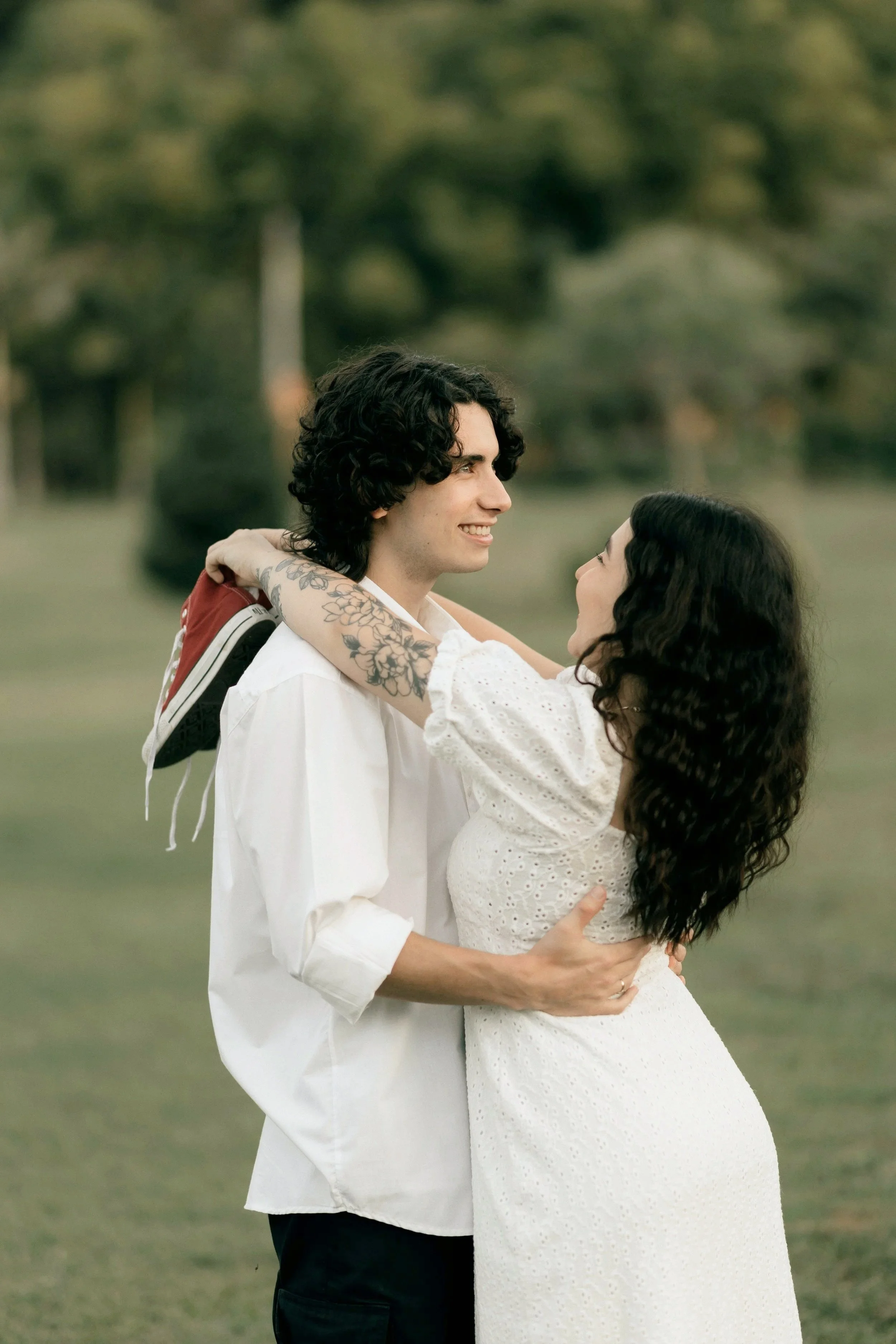A young man and woman are outside in a park, smiling and looking at each other. The woman is wearing a white dress and the man is wearing a white shirt and black pants. The man is holding the woman around her waist, and she has her arms around his neck.