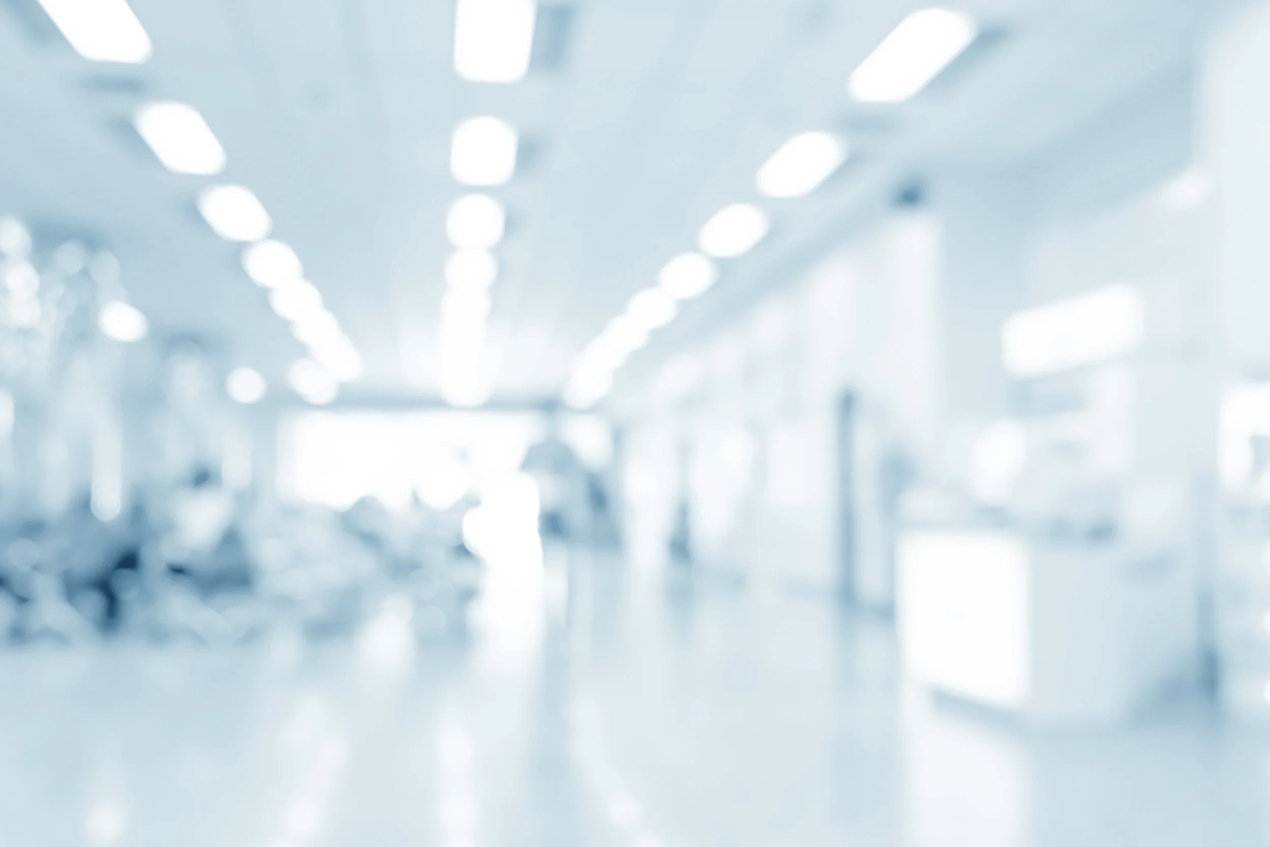 Blurred image of a hospital corridor with bright overhead lights and people walking in the distance.