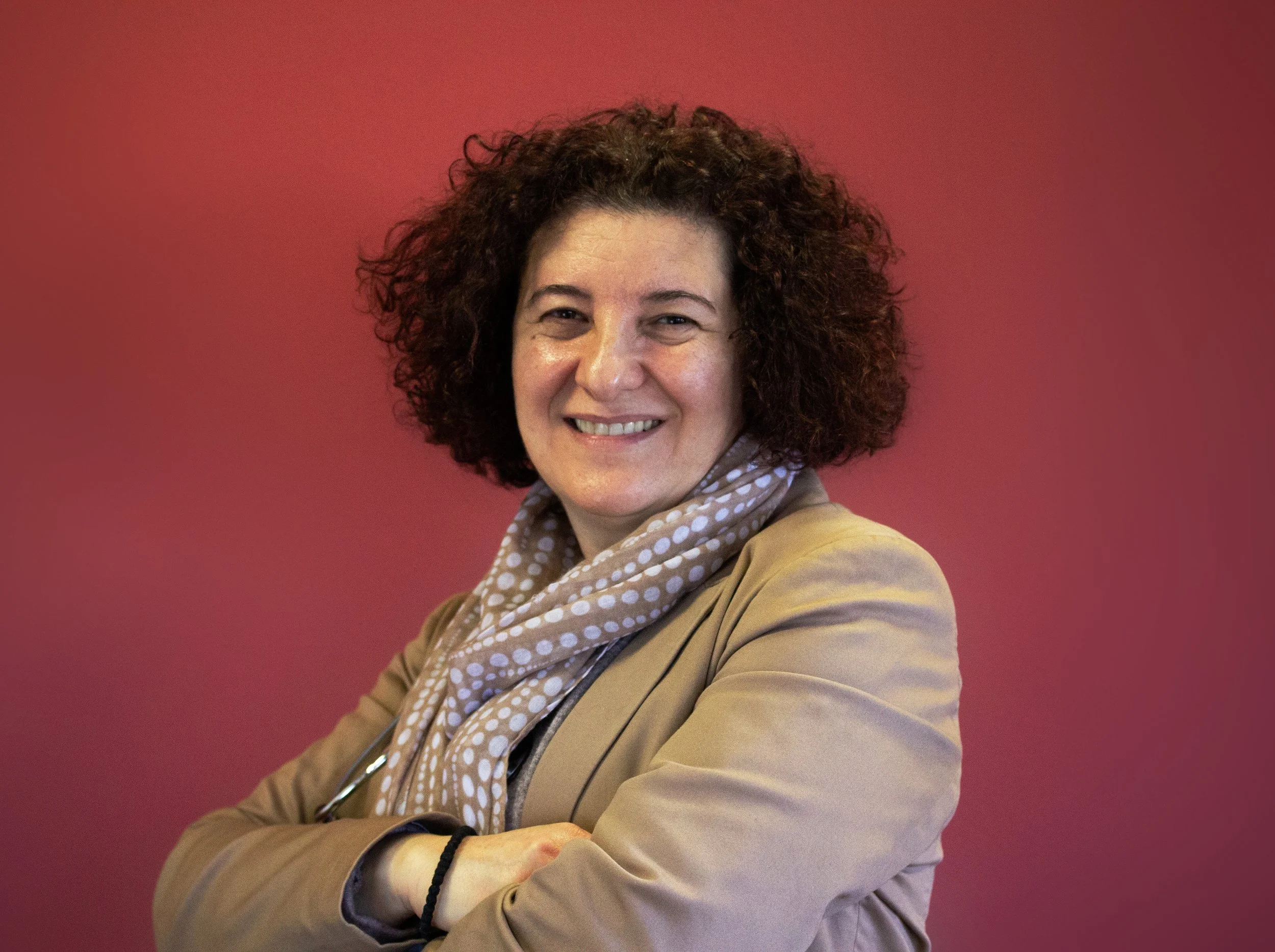 A woman with curly dark hair, smiling, wearing a beige blazer and a beige polka dot scarf, standing against a reddish background.