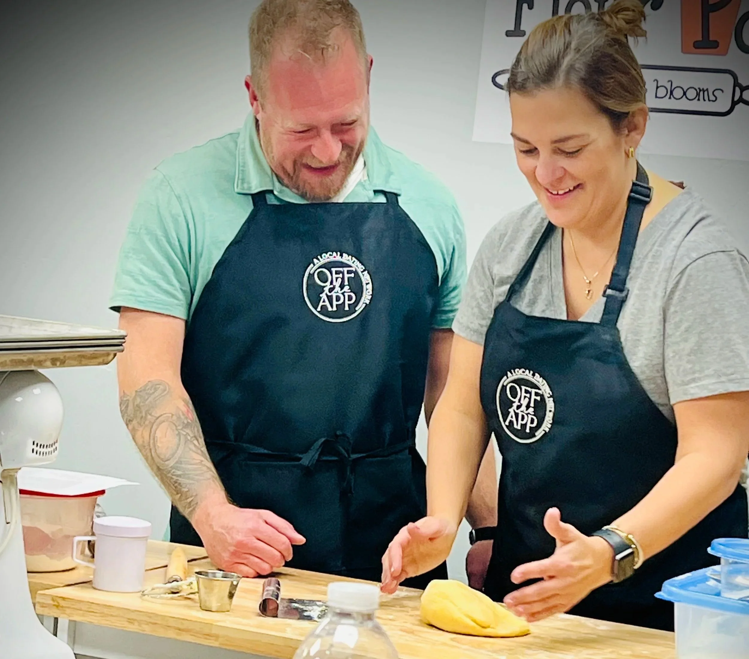 Two people wearing Off The App Dating aprons smiling while preparing food together at a hands-on singles event.