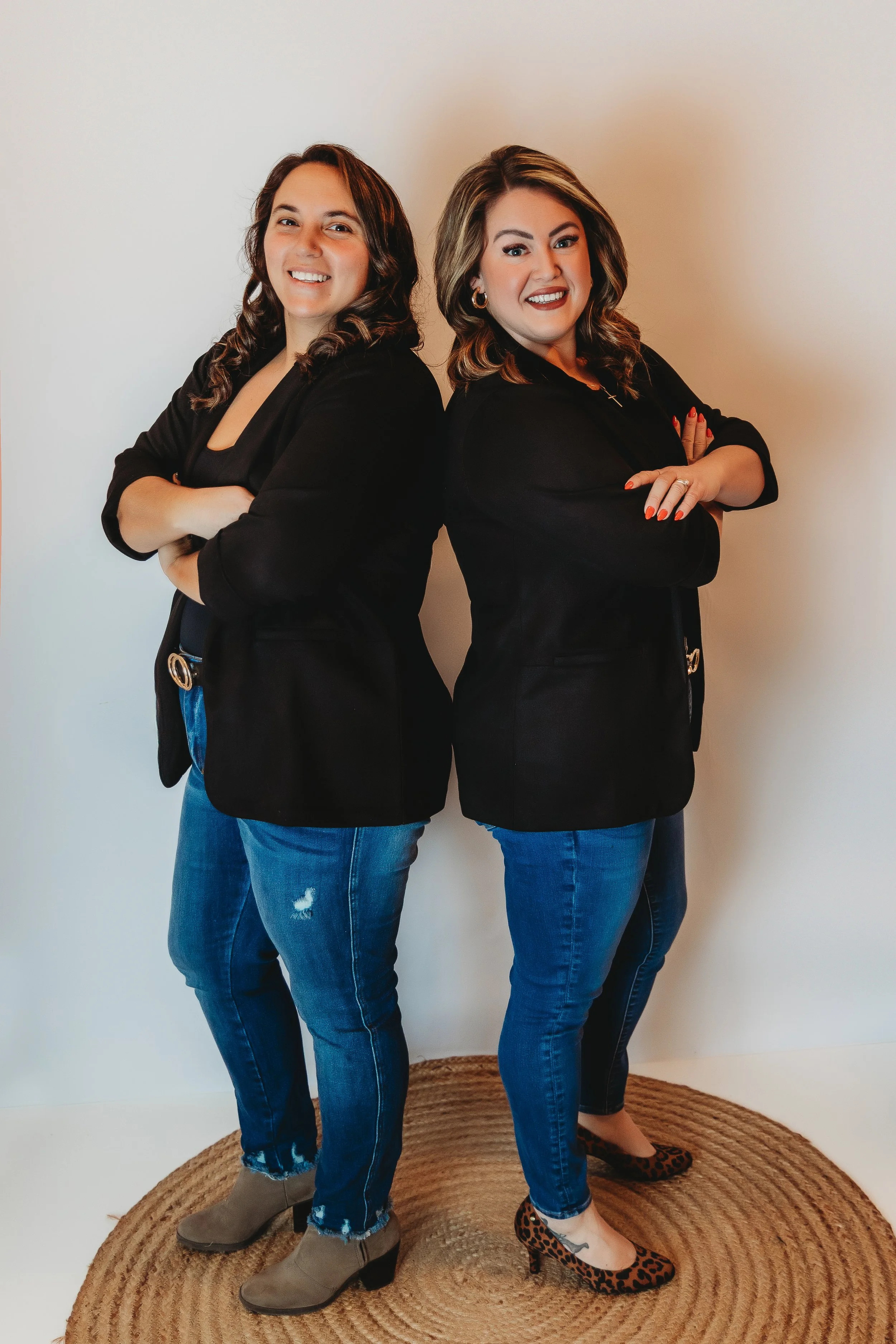 Brittany Smolenski and Cassandra Two women standing back to back, smiling confidently while wearing black tops and jeans in a studio setting