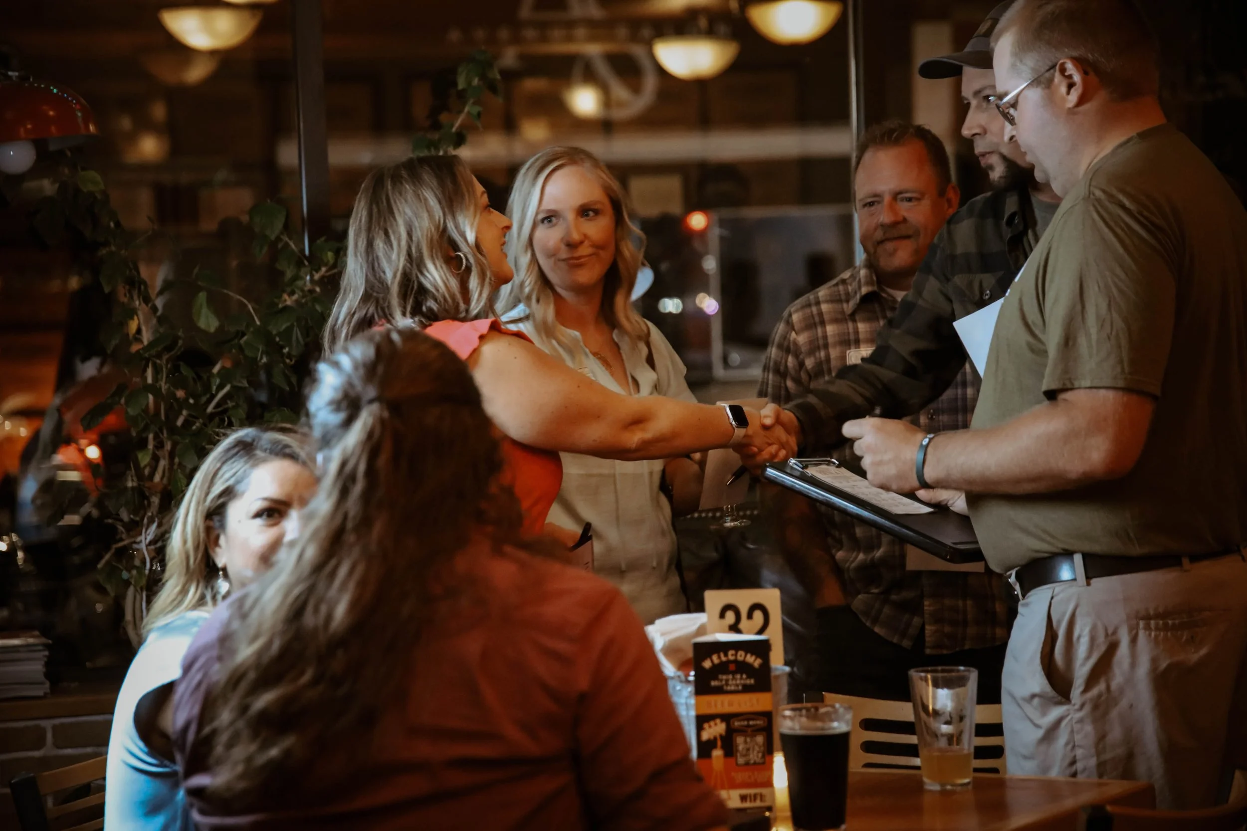 Group of singles gathered at a table while two people shake hands during an in-person dating event hosted at Appleton Beer Factory in Appleton, Wisconsin