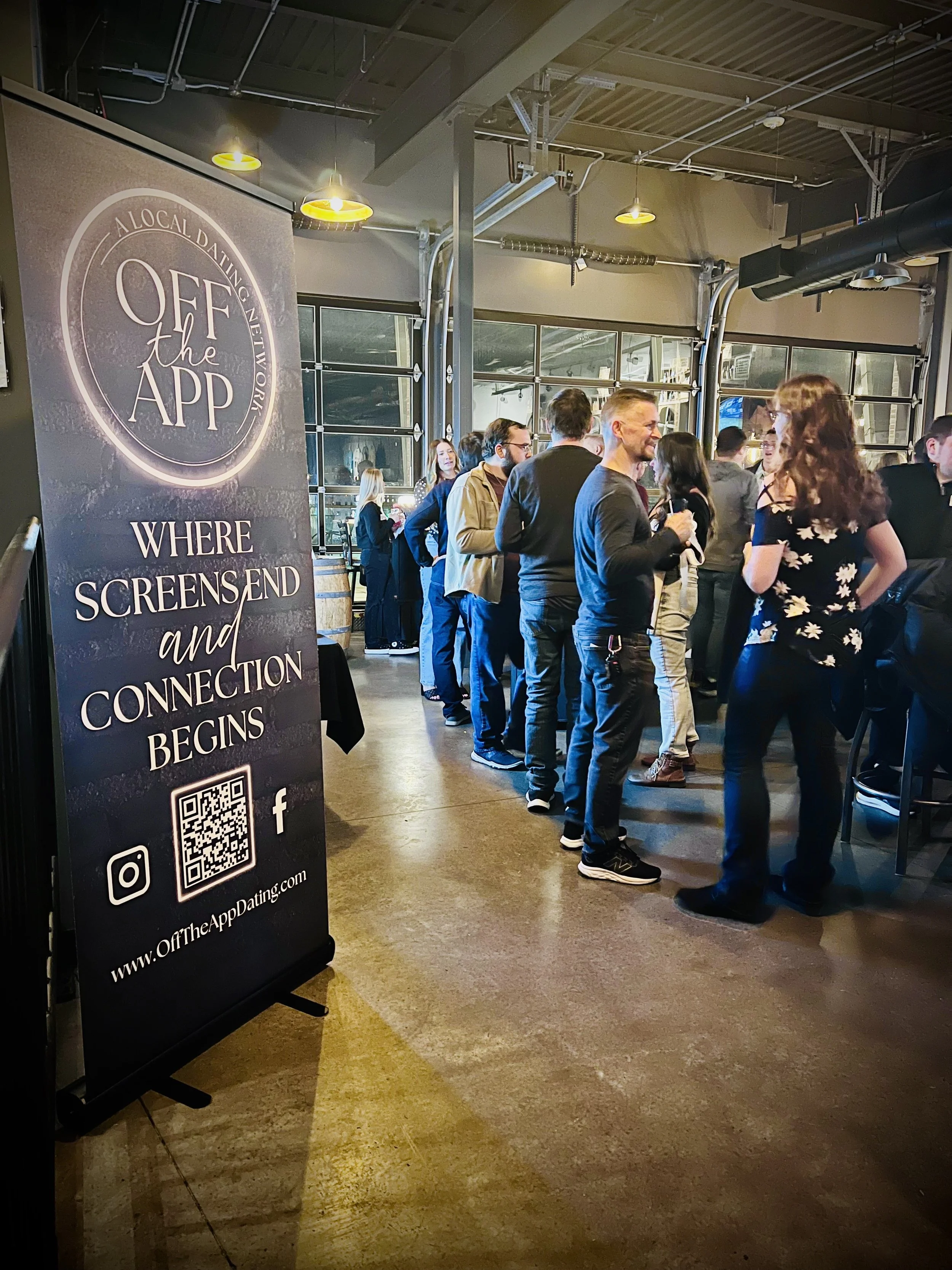 Attendees standing and socializing near an Off The App Dating banner at an in-person event hosted at Titletown Brewing Company in Green Bay, Wisconsin
