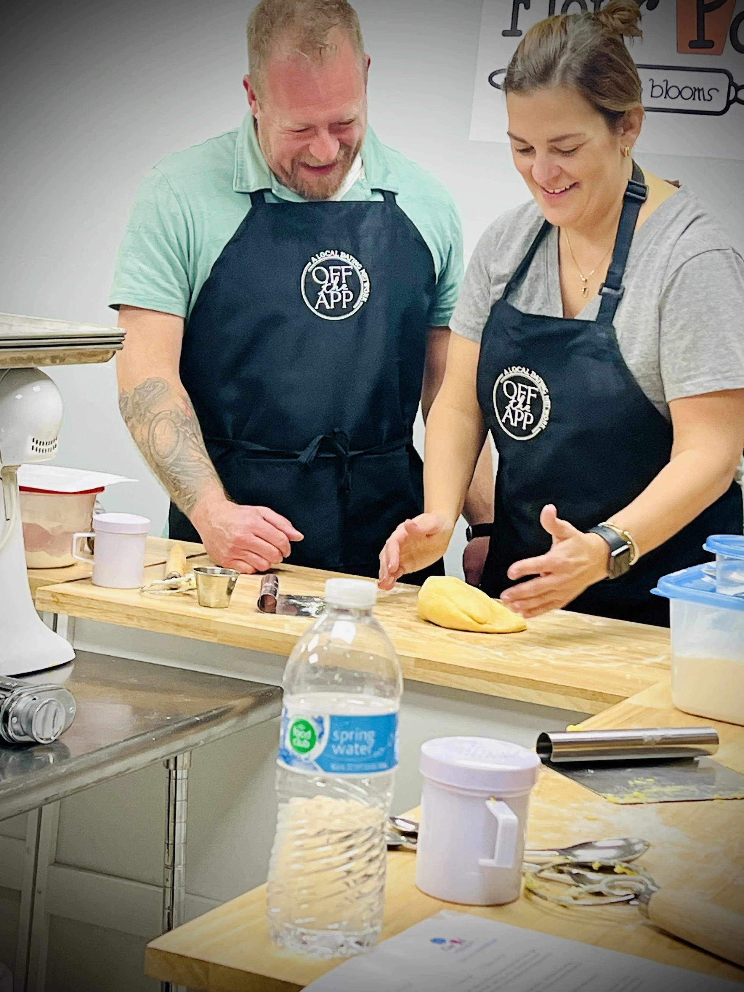 Two people wearing Off The App Dating aprons smiling while preparing food together at a hands-on singles event.