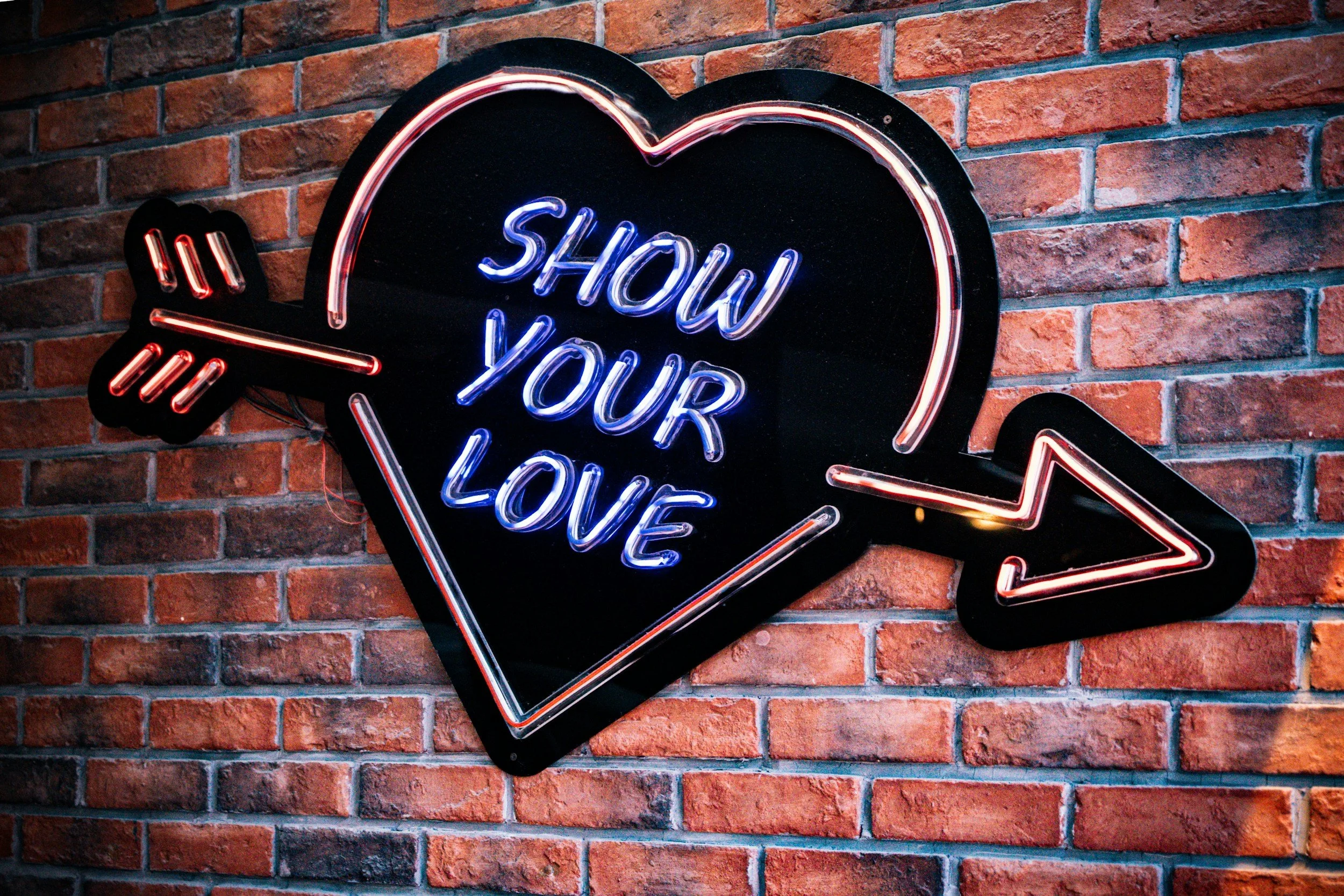 Neon sign shaped like a heart with an arrow through it, displaying the words "Show Your Love" in blue neon lights, mounted on a brick wall.