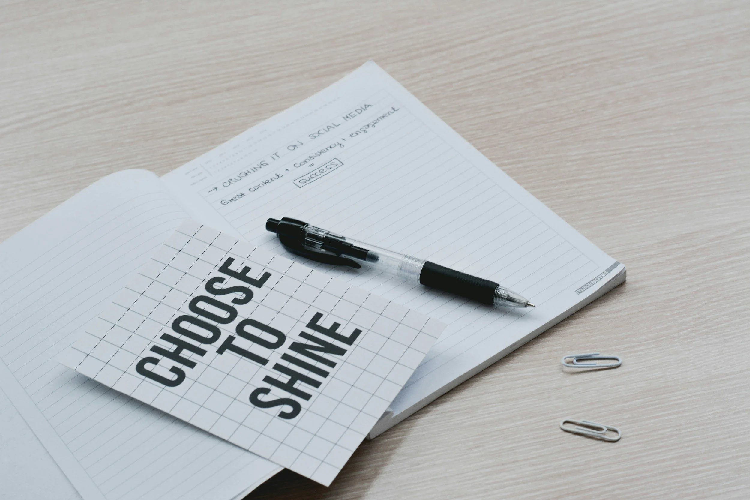 Open notebook with handwritten notes, a black pen, and paper clips on a light wooden table.