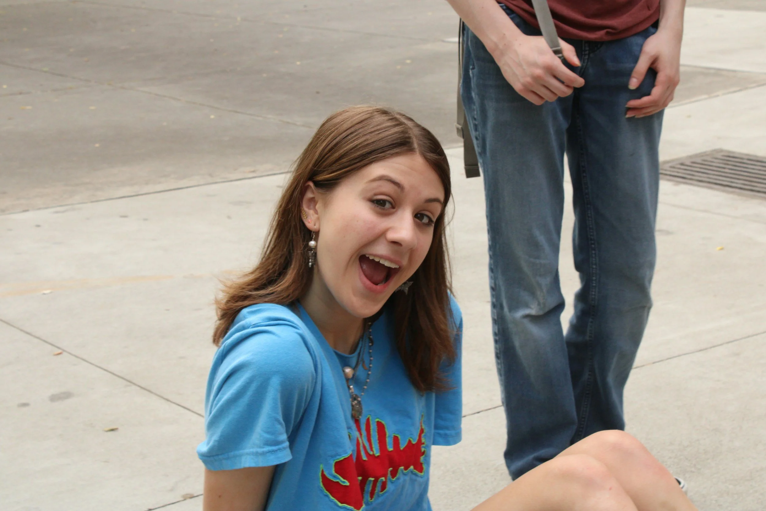 A young woman with brown hair and earrings is smiling and looking at the camera. She is wearing a blue shirt and is outdoors on a paved surface. Partially visible is a person standing beside her in jeans and a maroon shirt.