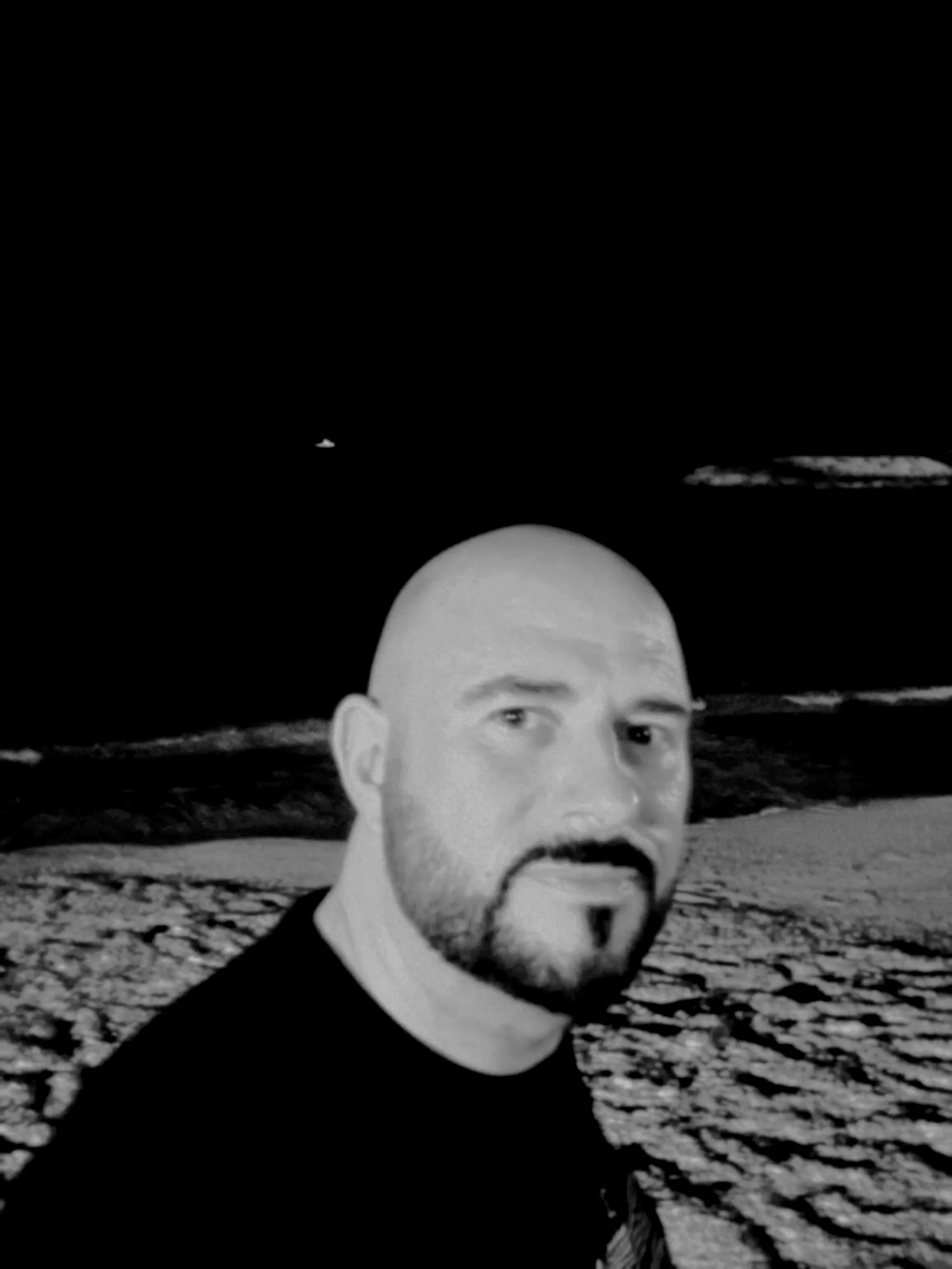 Black and white photo of a man with a bald head, beard, and mustache, standing on a beach at night with the moon visible in the sky.