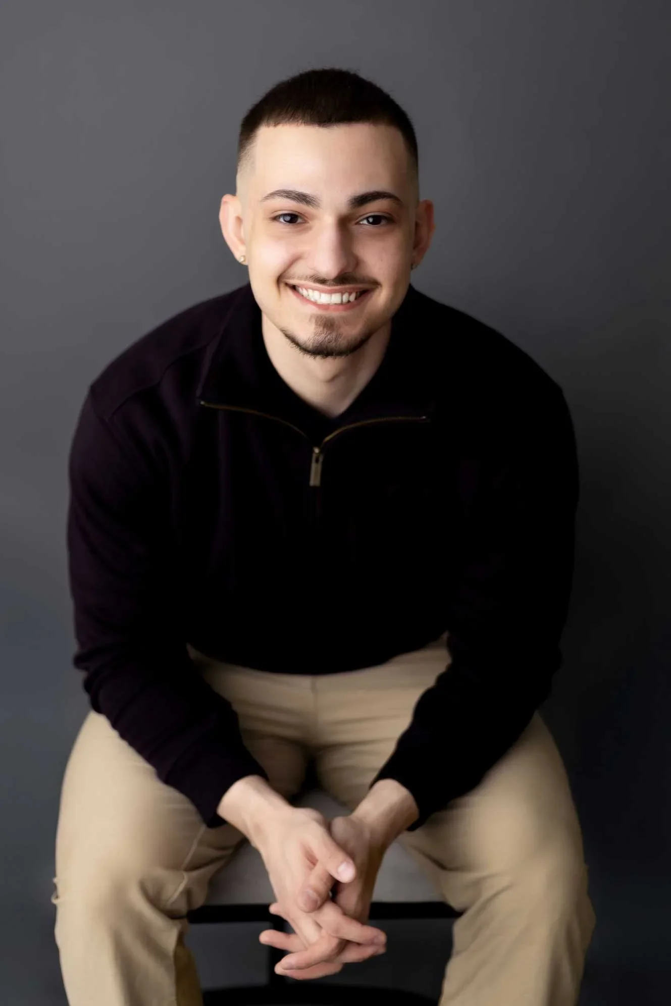 A young man with a short beard and mustache, smiling, wearing a black zip-up sweater and beige pants, sitting against a plain gray background.