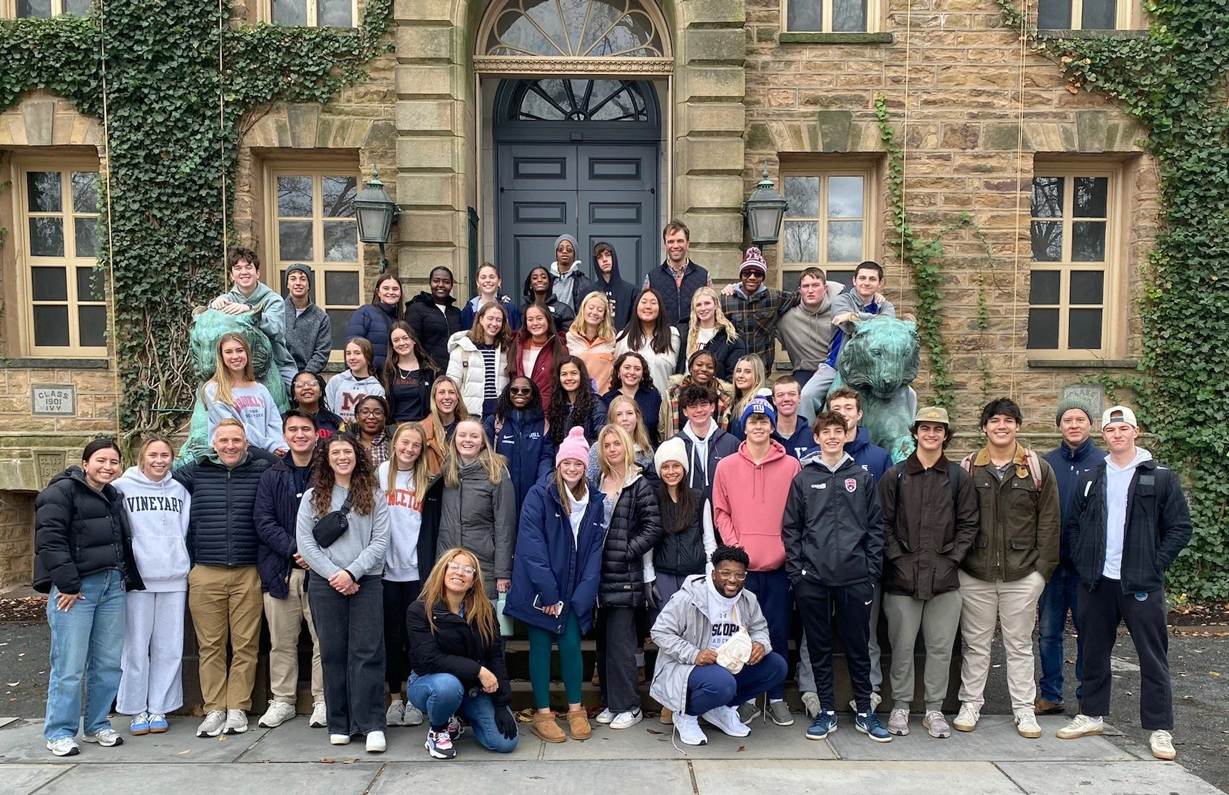 A large group of diverse young students and adults poses for a photo on steps outside a historic stone building with ivy-covered walls and dark blue door, and lion statues.