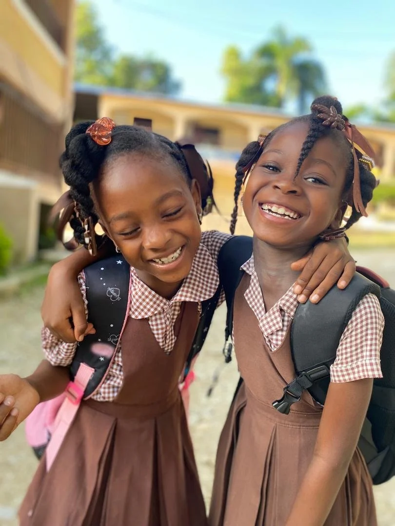Two young girls in school uniforms hugging and smiling outdoors, wearing backpacks.